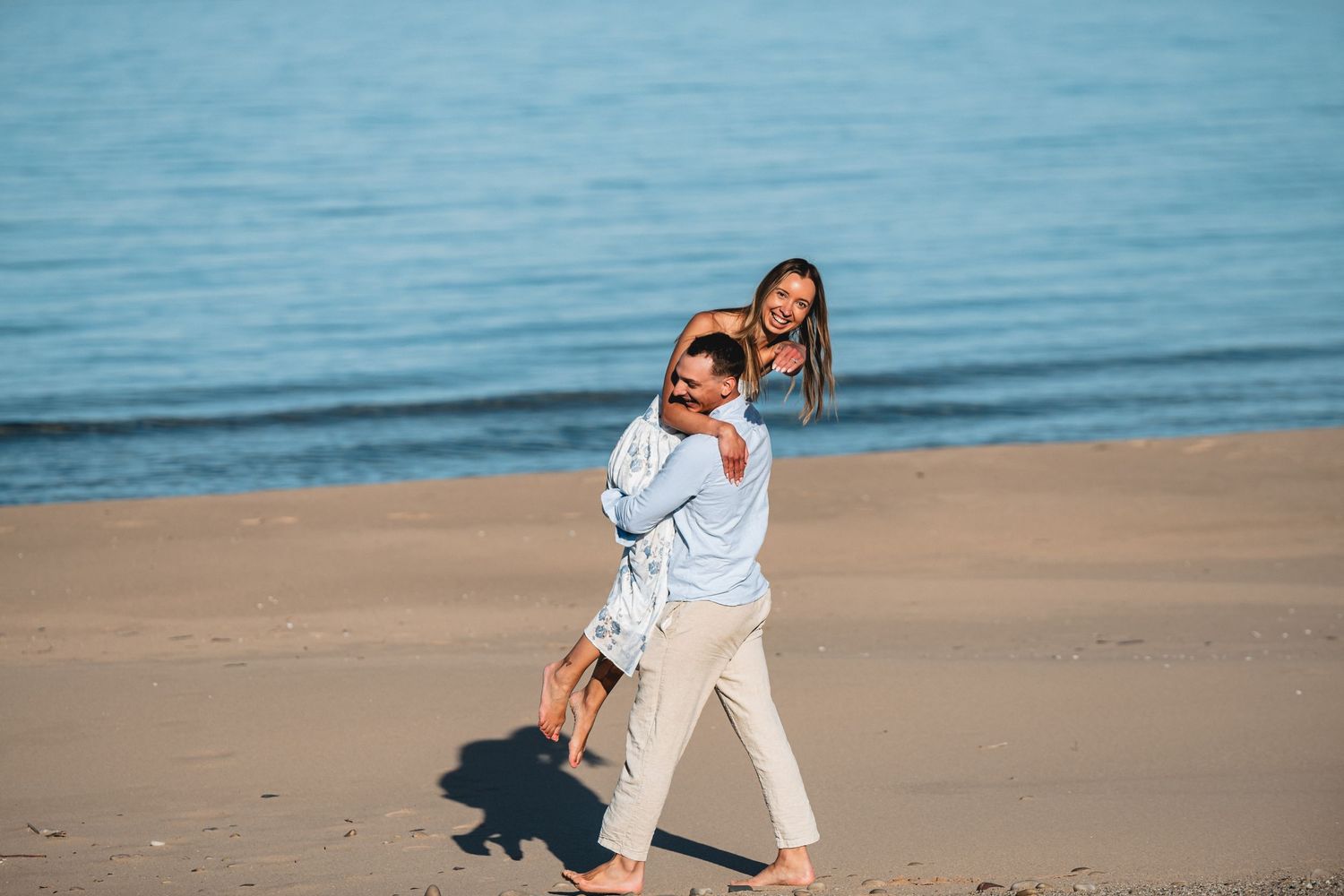 A happy couple enjoys a romantic piggyback ride on a sunny sandy beach with calm blue ocean water in the background.