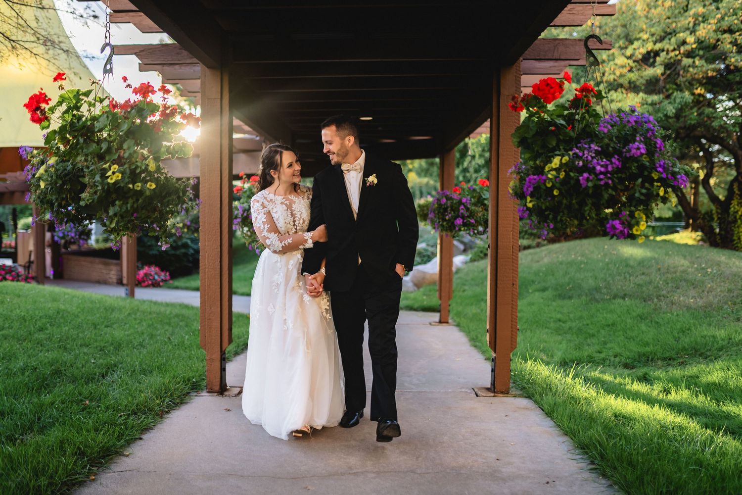 A couple walks together in a garden setting with beautiful flowering plants and trees along a covered walkway.