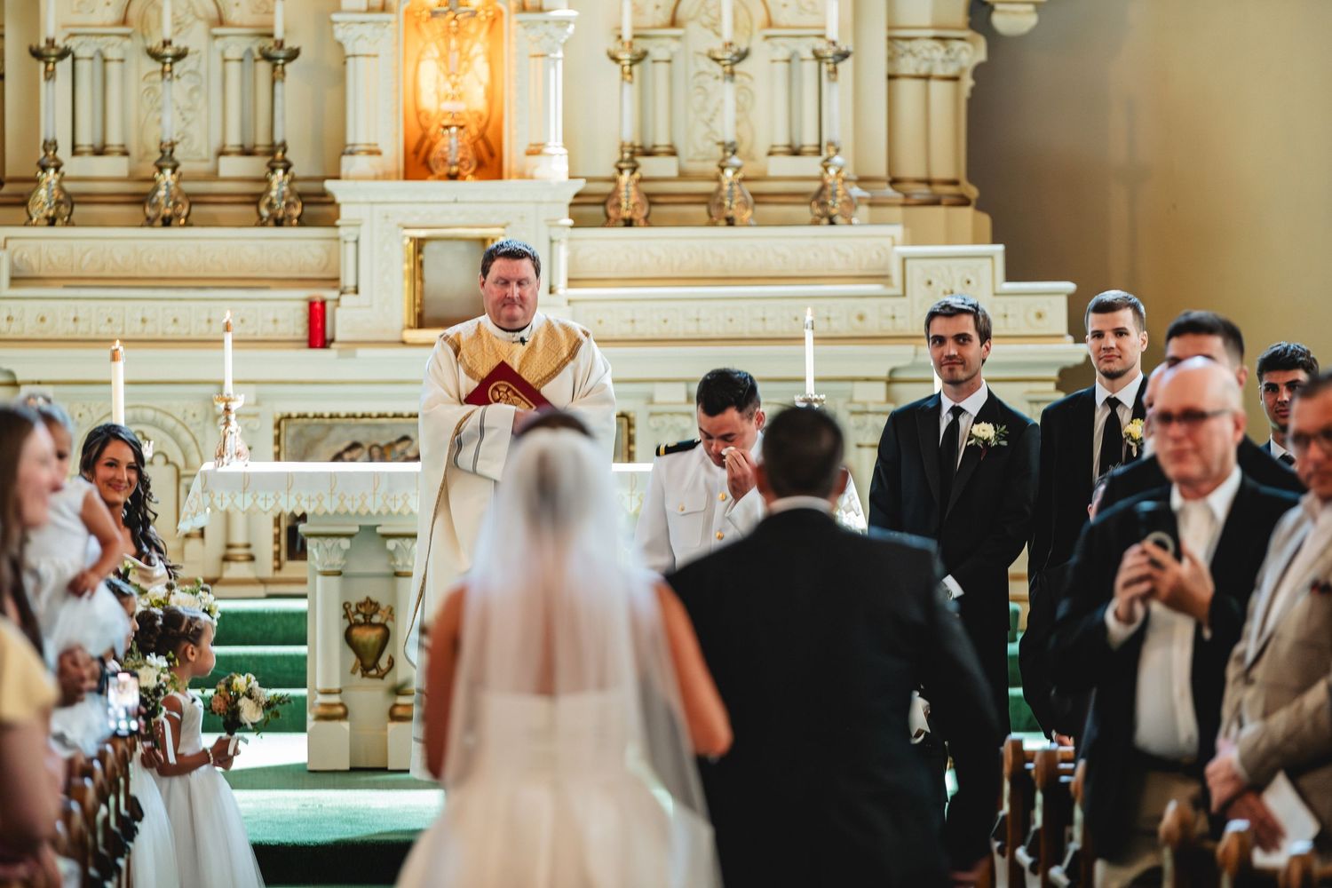 Wedding ceremony in an ornate church with white columns and chandeliers as guests watch the couple exchange vows.