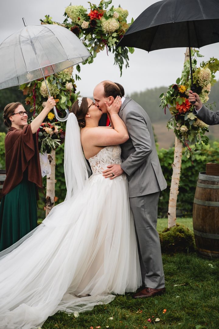 Wedding couple shares romantic kiss under clear umbrellas in garden setting with floral arch and wine barrels.