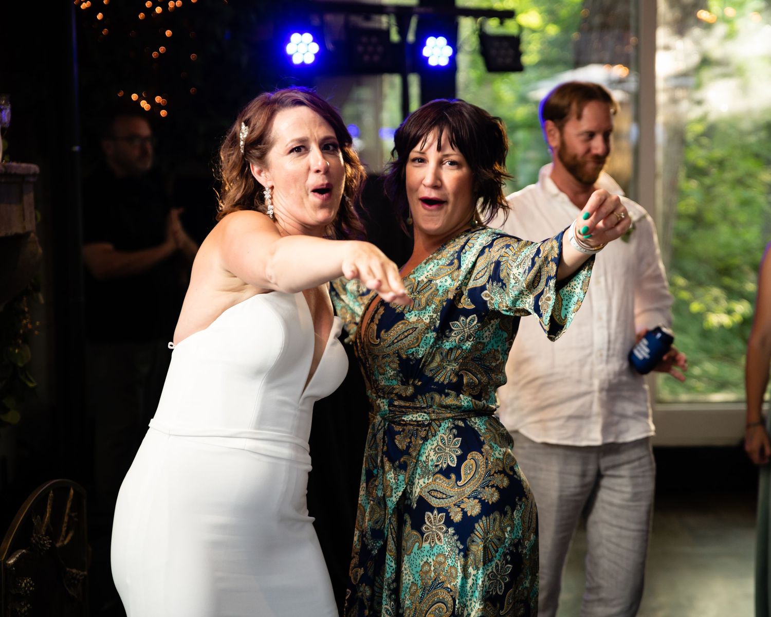 Wedding guests dancing together in a reception hall with festive lighting and greenery in the background.