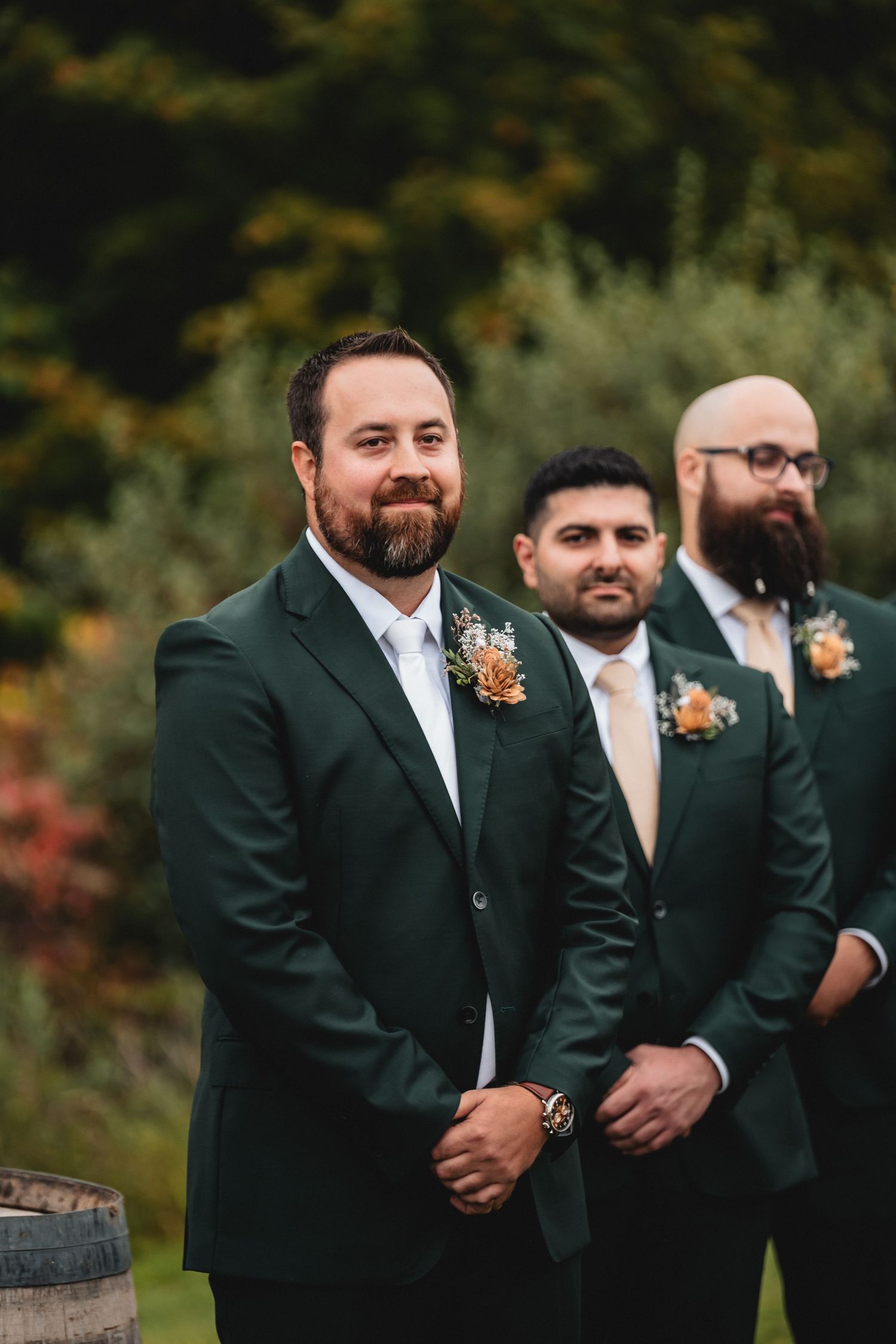Wedding party members in dark suits with orange and white floral boutonnieres stand together outdoors in autumn.