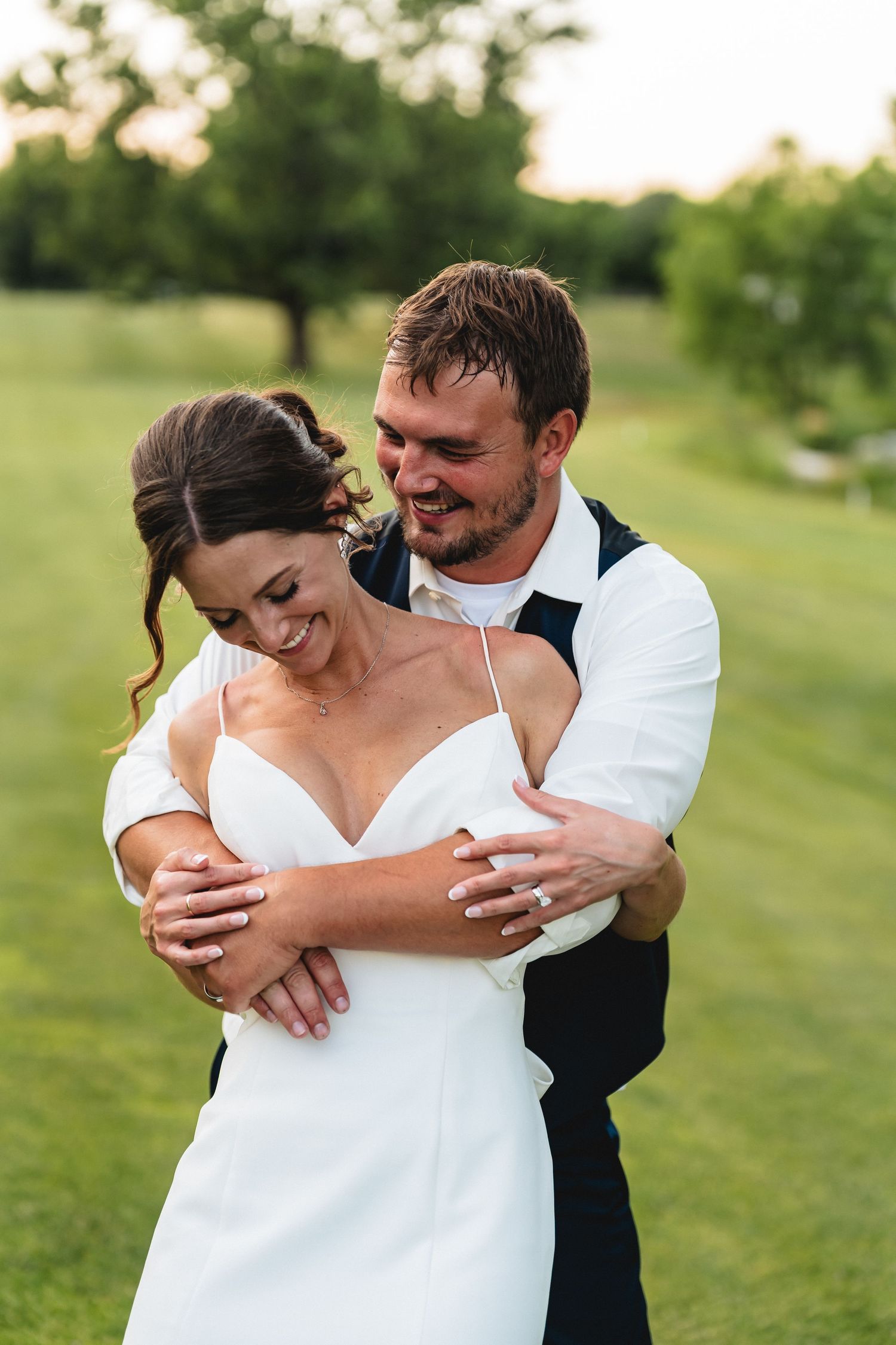 Bride and groom embracing outdoors on green lawn at golden hour, smiling joyfully in wedding attire.