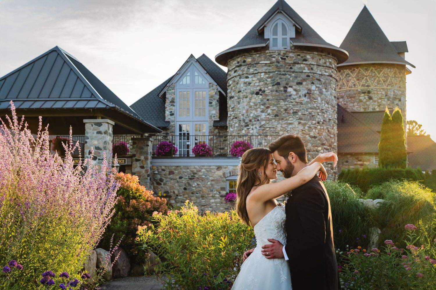 A romantic wedding couple embraces at sunset in front of a rustic stone building surrounded by blooming gardens.