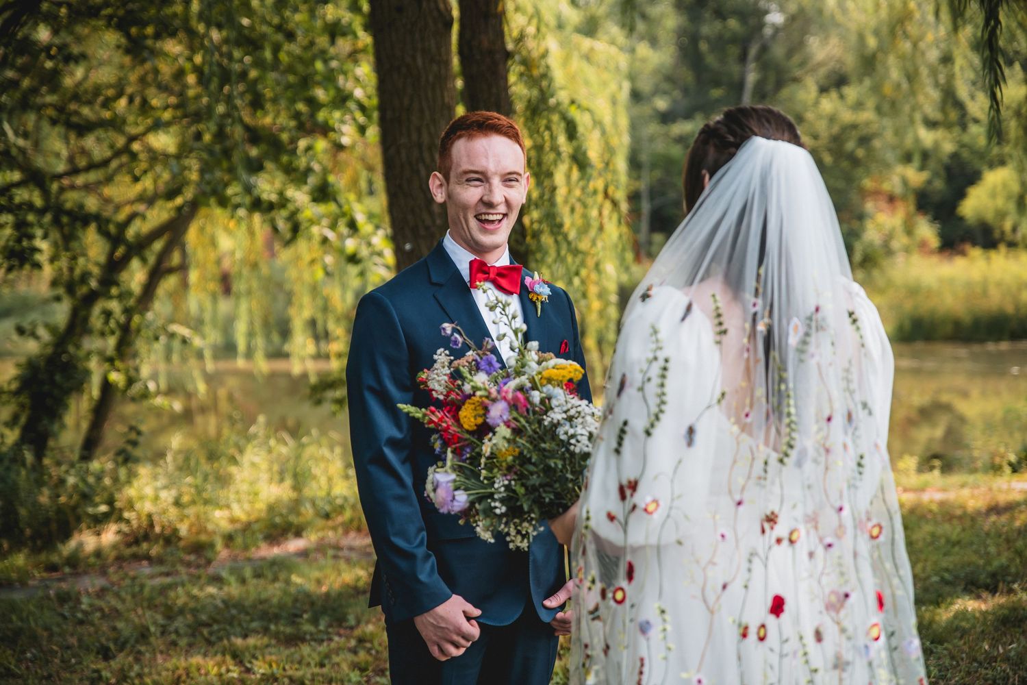 Smiling groom in blue suit holds wildflower bouquet beside bride in floral embroidered veil outdoors.