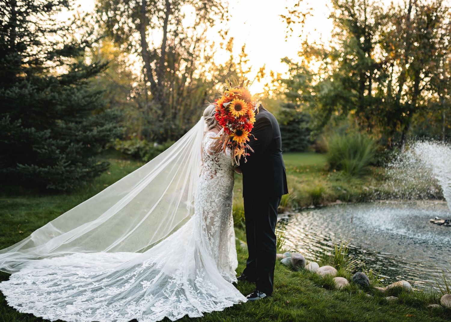 Bride and groom kissing behind sunflower bouquet in lace wedding gown with veil by garden pond at sunset.