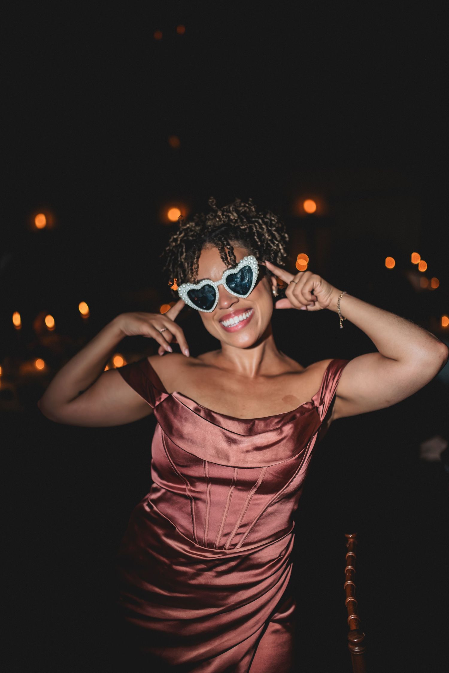 Person in pink satin dress and white sunglasses poses playfully against dark background with glowing orange lights.