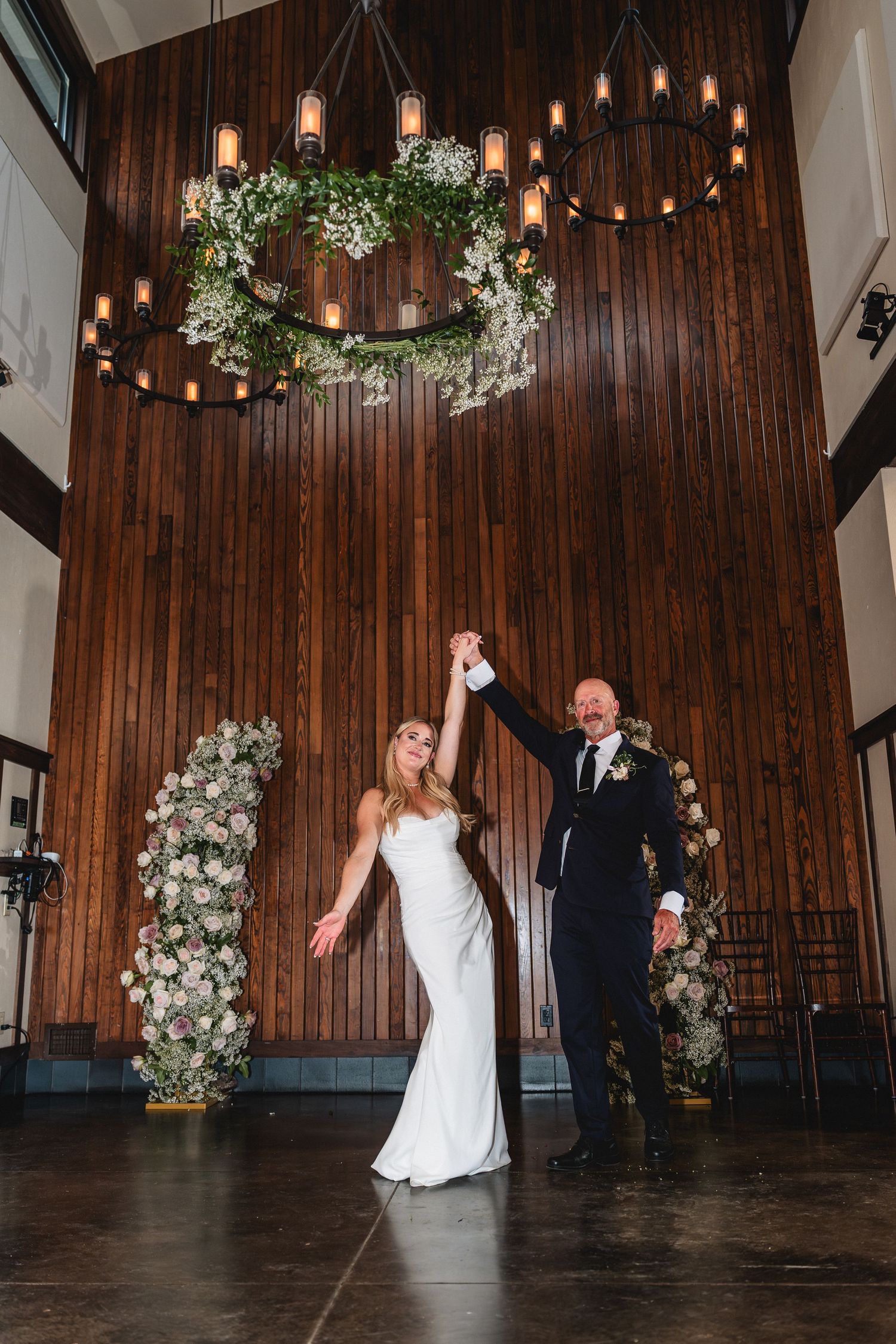 Bride and groom share first dance under floral chandelier in elegant wood-paneled wedding venue with candle lighting.