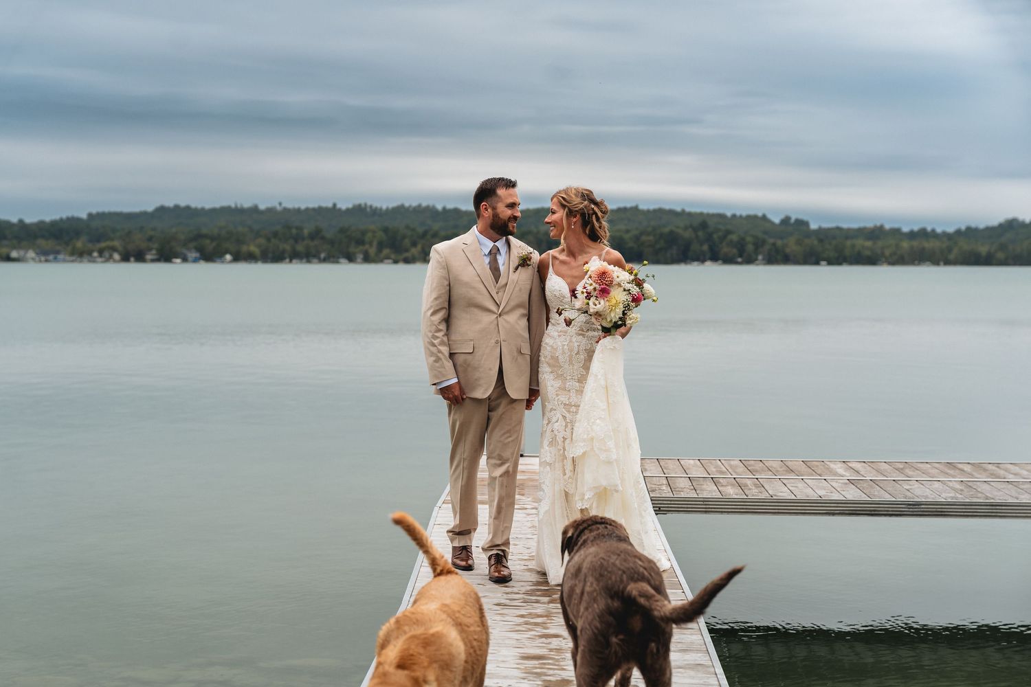 Bride and groom stand on a lakeside dock with two dogs, surrounded by serene water and lush green trees captured in Traverse City