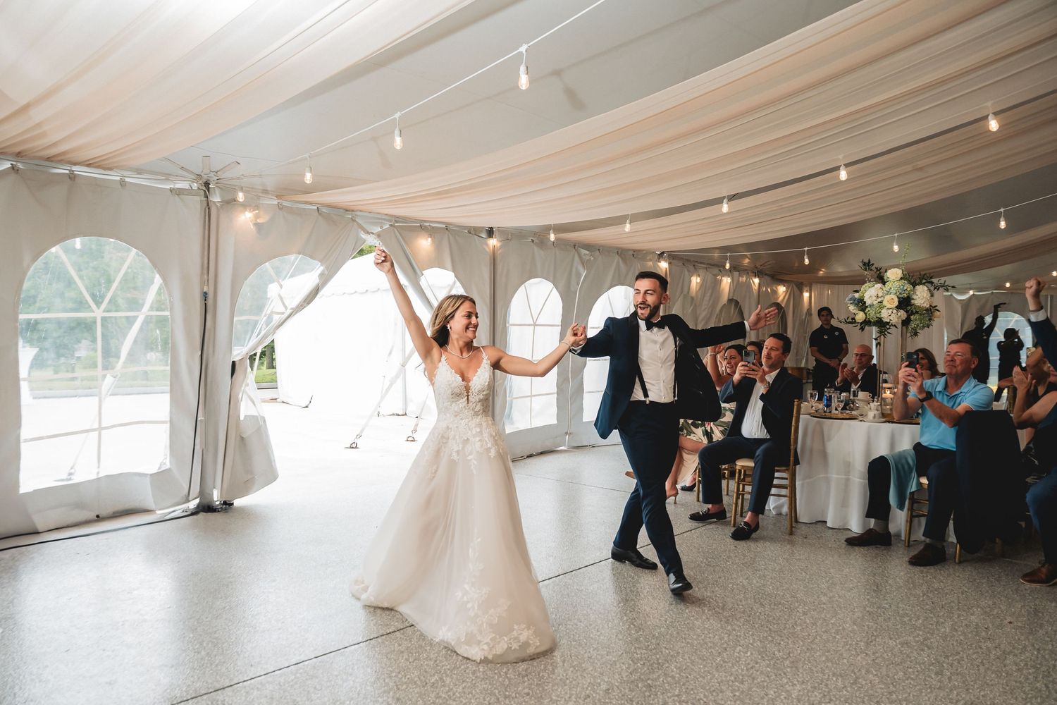 Bride and groom share their first dance under string lights in an elegant white draped wedding reception tent.