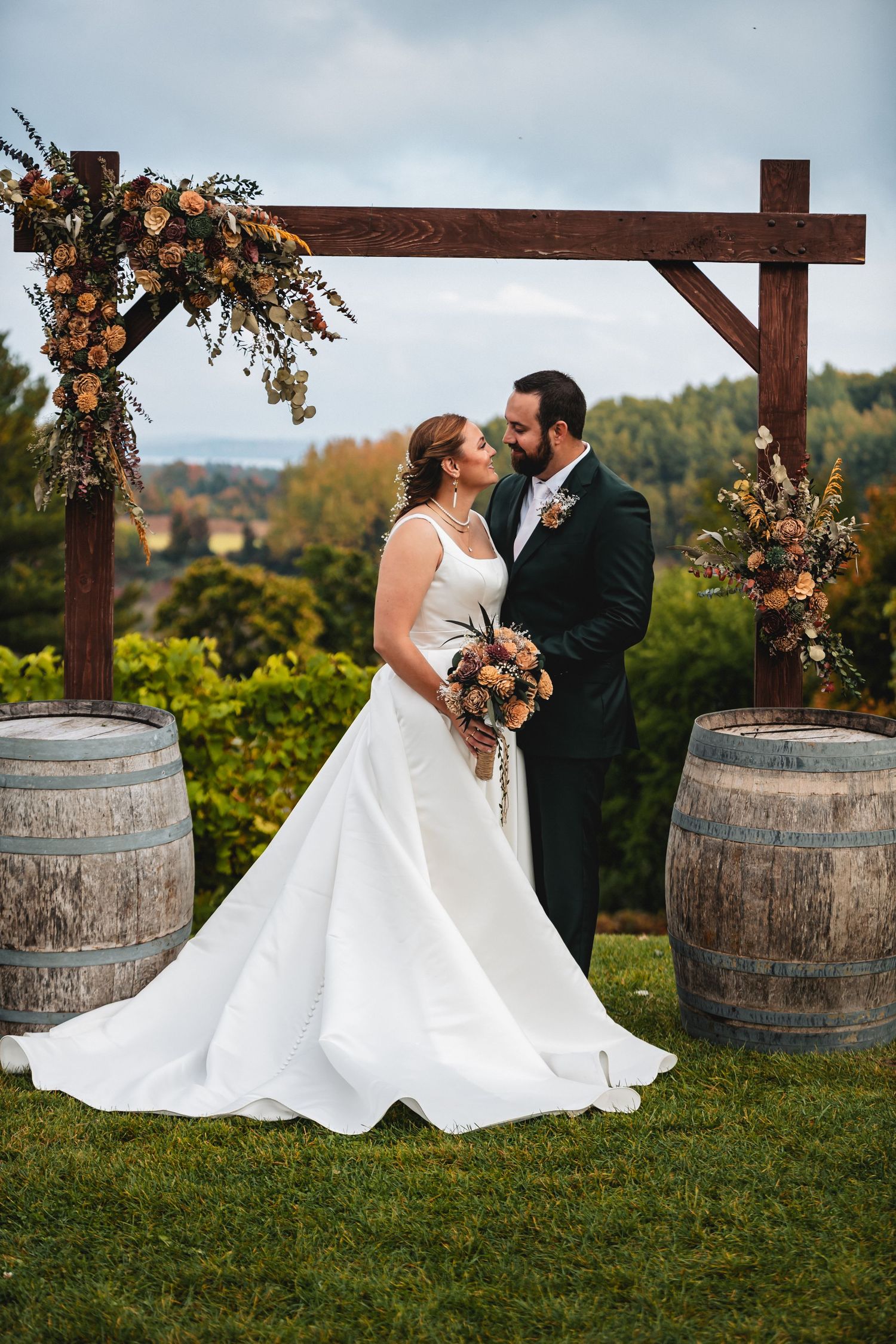 Romantic wedding couple poses near rustic wine barrels and wooden arch decorated with autumn florals at outdoor vineyard.