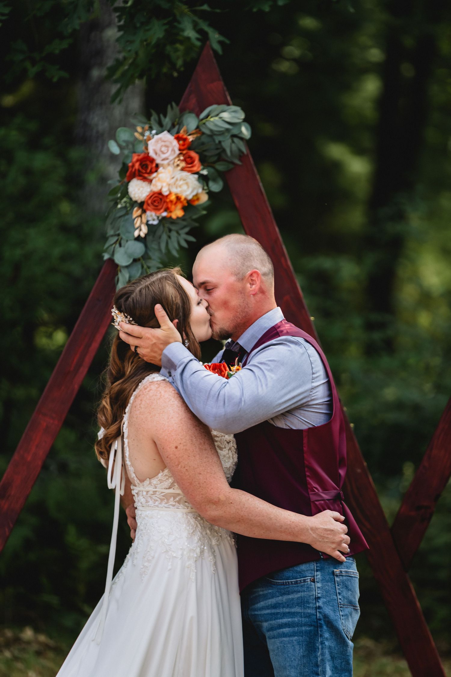 Bride and groom share first kiss under rustic triangular wooden arch with fall floral arrangement in forest setting.
