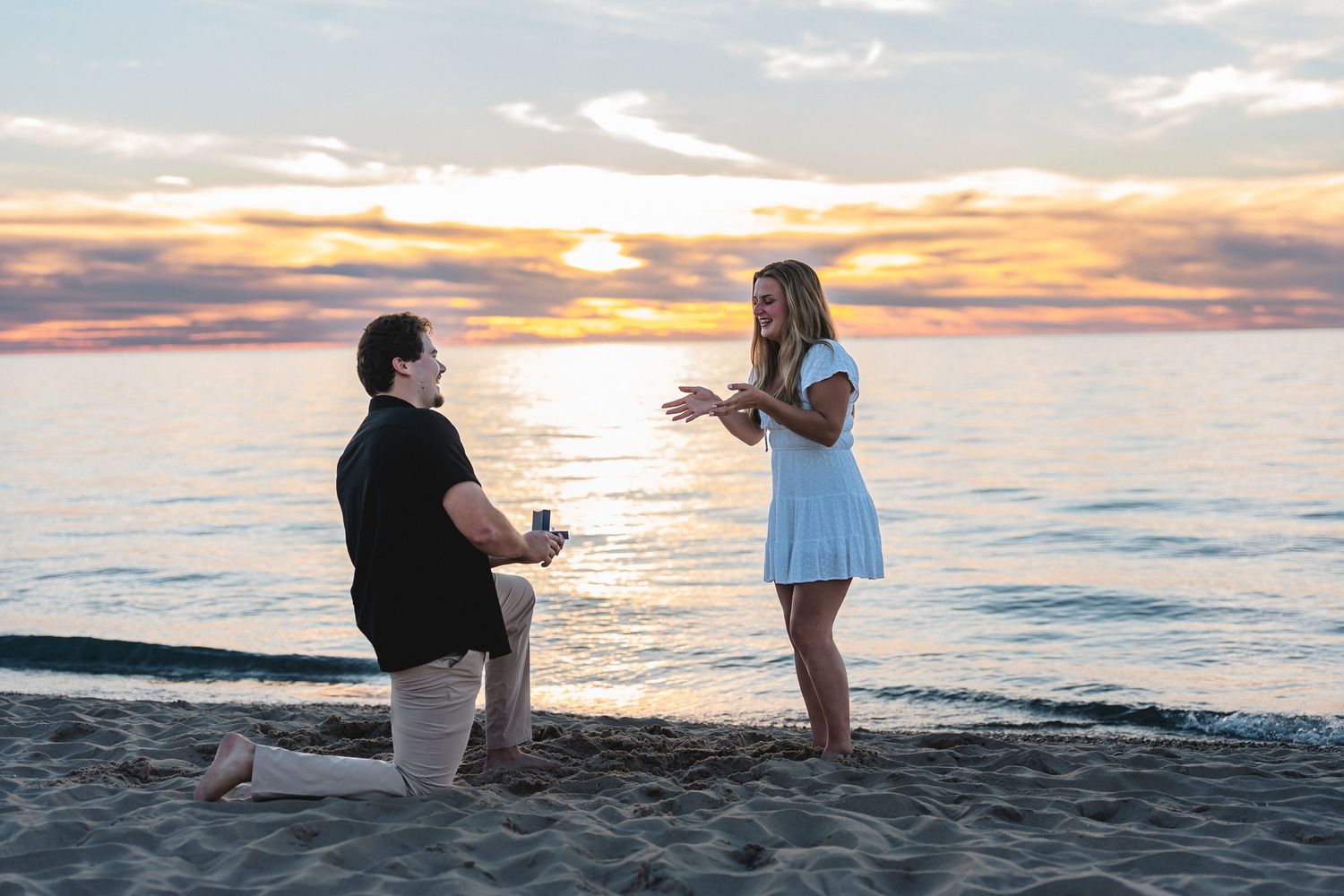 Man kneeling on one knee proposes to a smiling woman in a white dress on a sandy beach near Frankfort, MI at sunset.