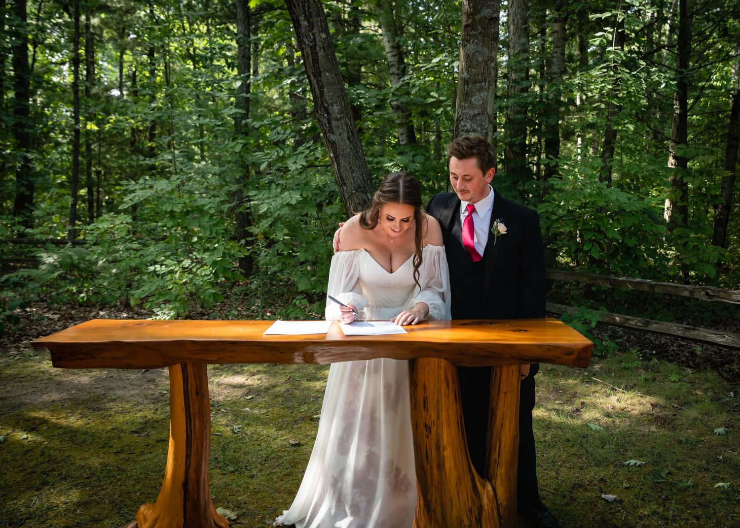 A couple in wedding attire sits together at a rustic wooden table in a lush forest setting.