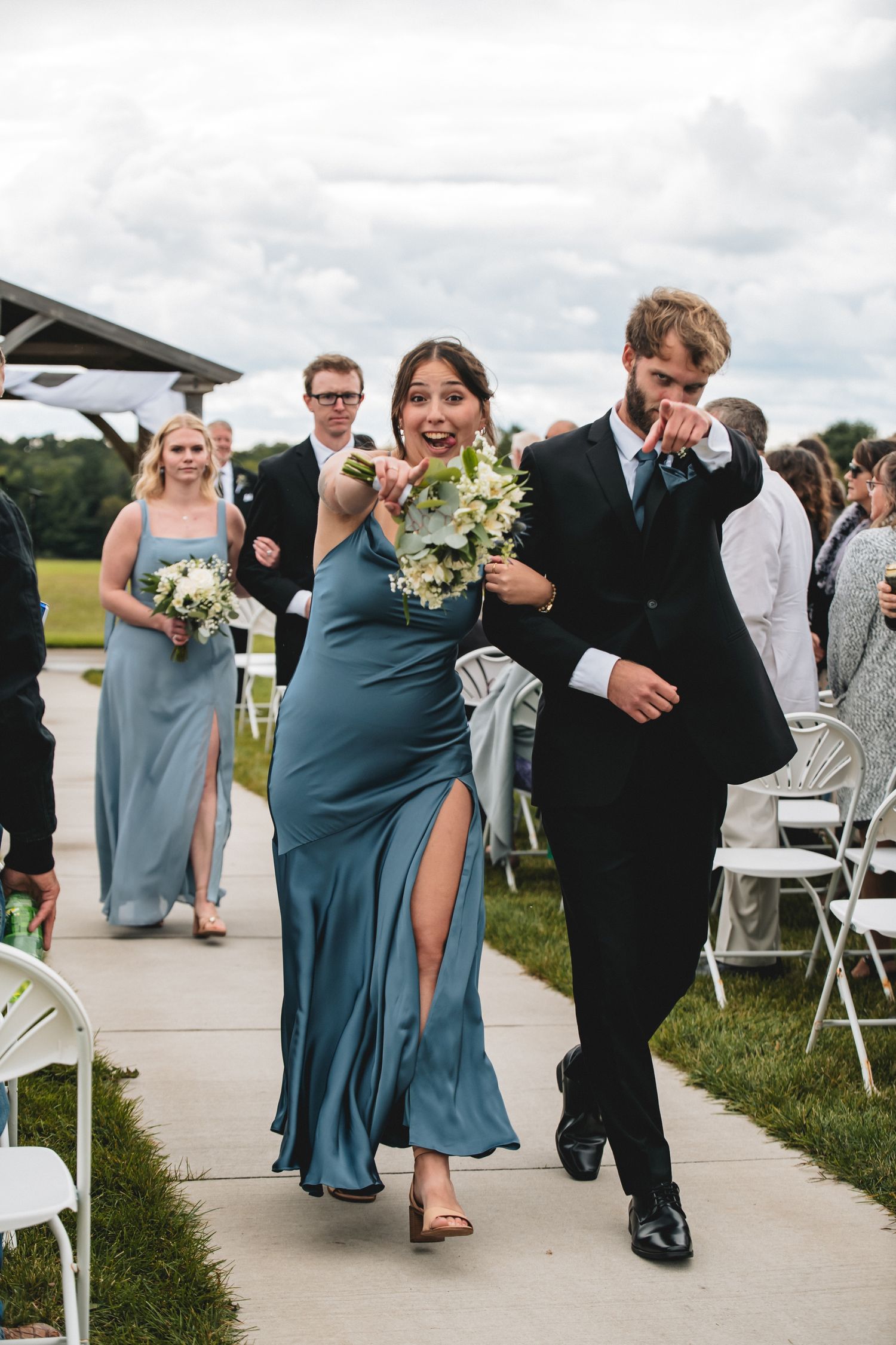 Wedding party members in blue dresses and black suits walk and laugh together on a paved outdoor path after ceremony.