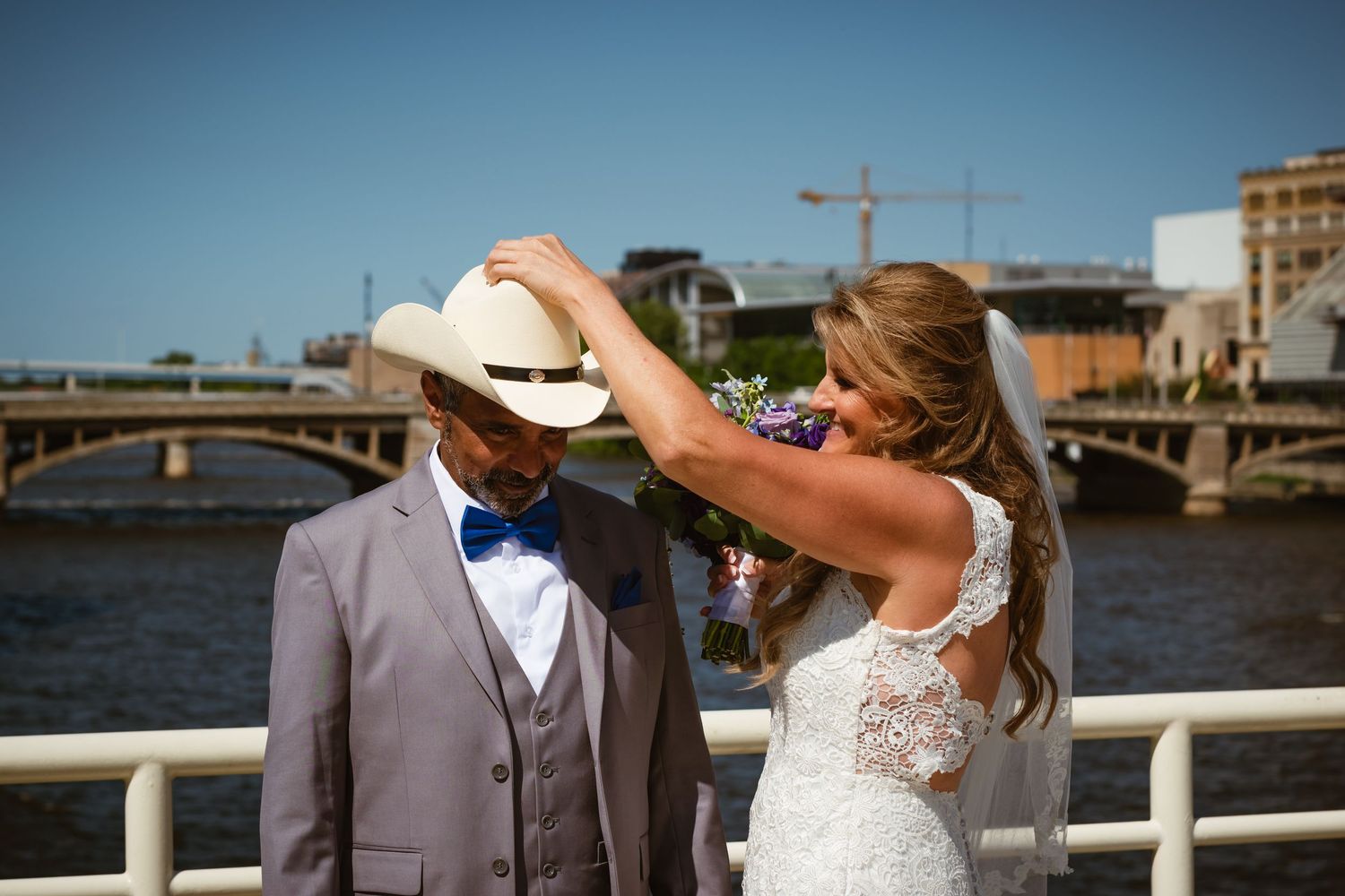 A bride adjusts her grooms cowboy hat during wedding portraits at the Blue Bridge in front of a Grand Rapids scenic river backdrop.
