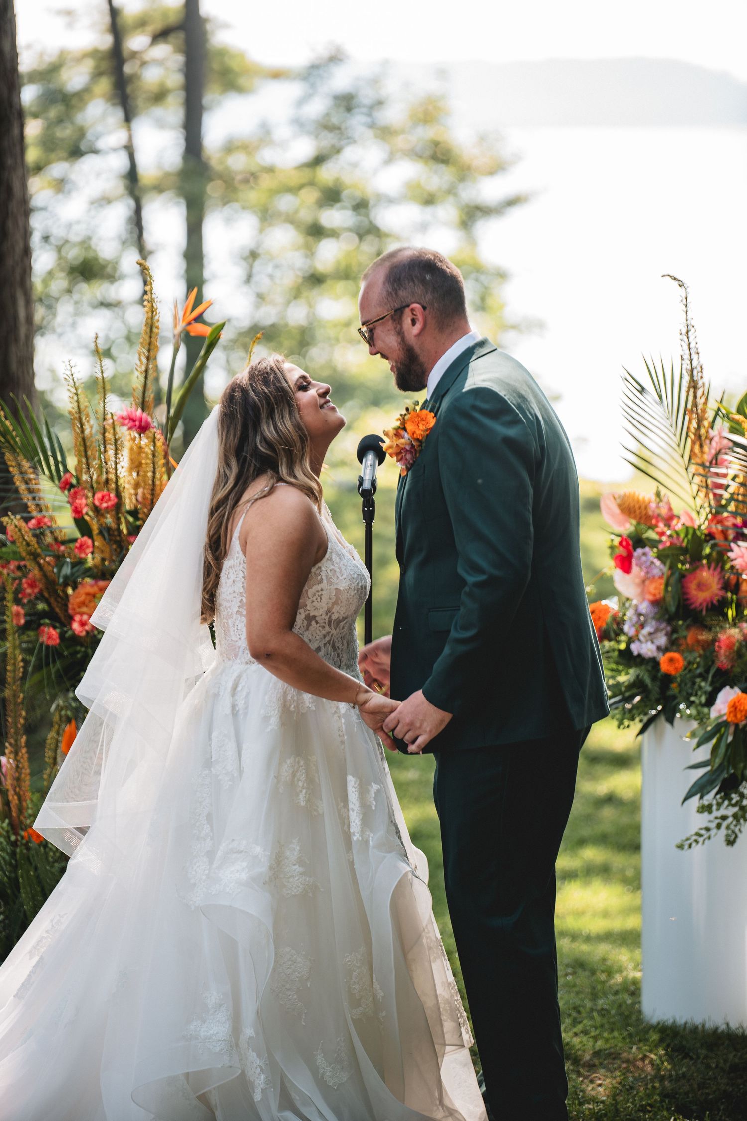 A romantic outdoor wedding ceremony features bright orange and pink tropical floral arrangements against a natural backdrop.