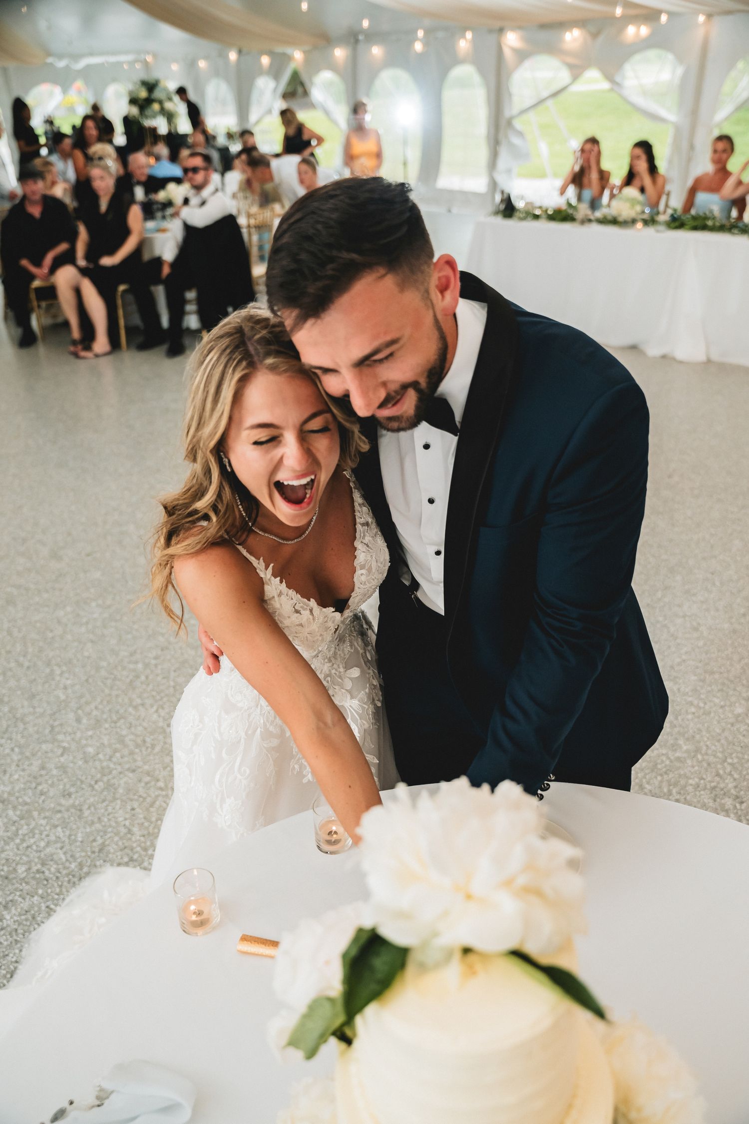 Joyful bride and groom cutting white wedding cake together at elegant tented reception, laughing and smiling.