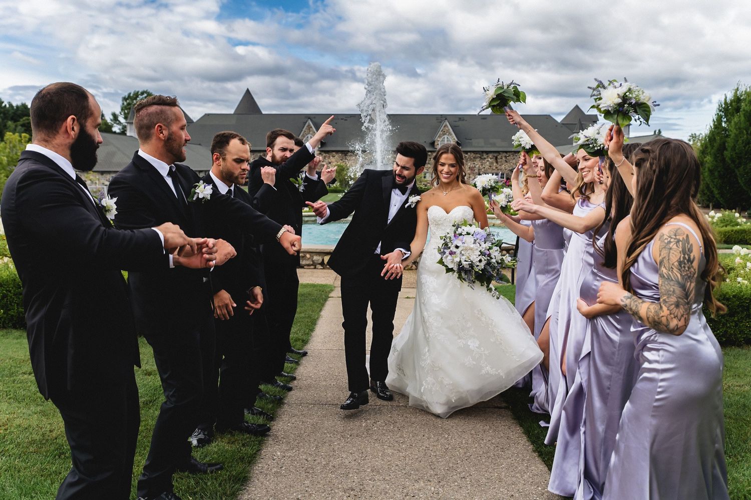 Bride and groom walk through cheering wedding party with groomsmen and bridesmaids in lavender gowns by fountain.
