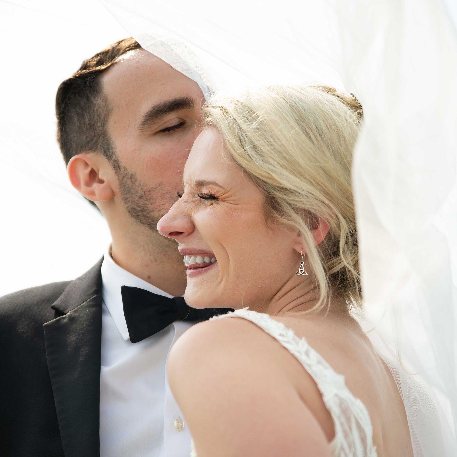 A groom in a black tuxedo kisses the cheek of a smiling bride in a white gown and veil on their wedding day.