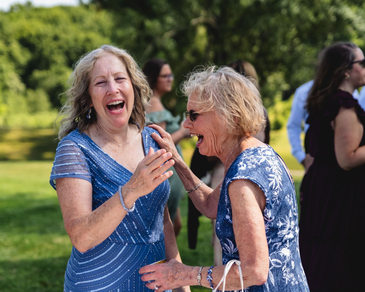 Two women in blue dresses laughing joyfully outdoors at a garden party event.