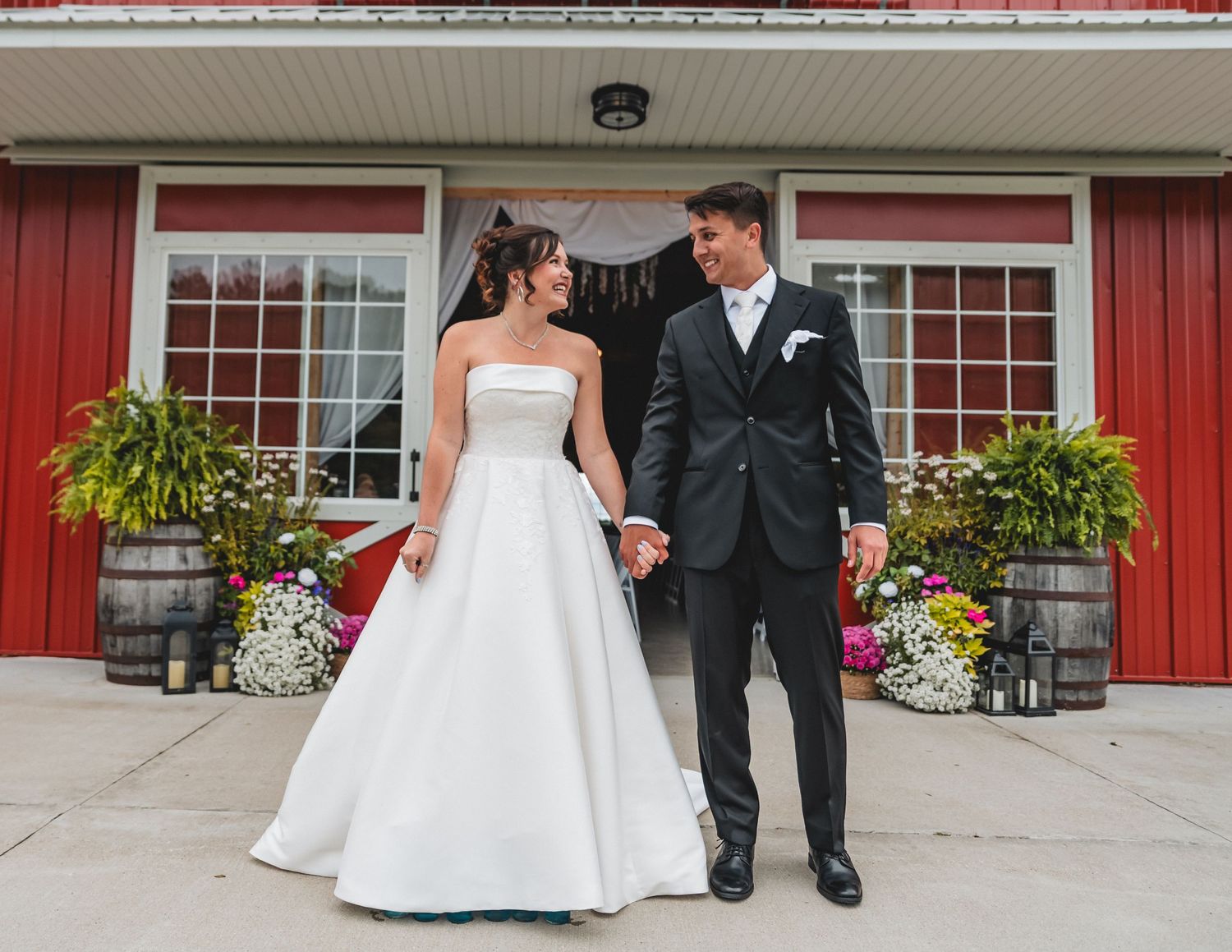 Bride and groom holding hands smiling at each other outside a red barn wedding venue with floral decorations.