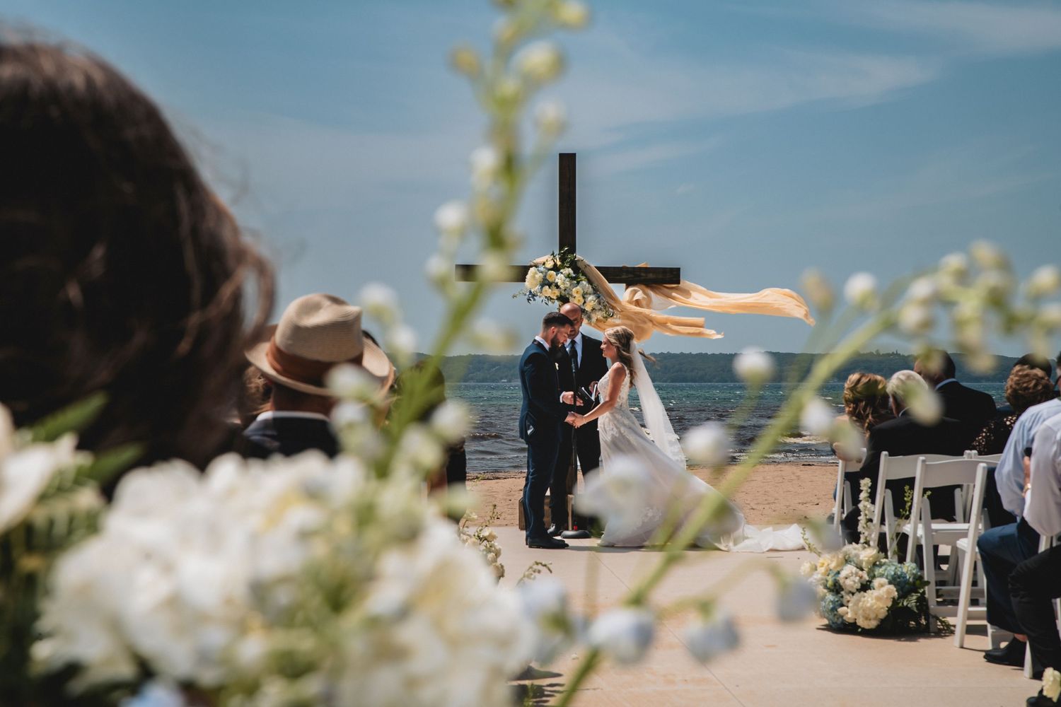 Outdoor beach wedding ceremony with couple at floral arch, cross backdrop, ocean view, white chairs, and guests seated on sunny day.