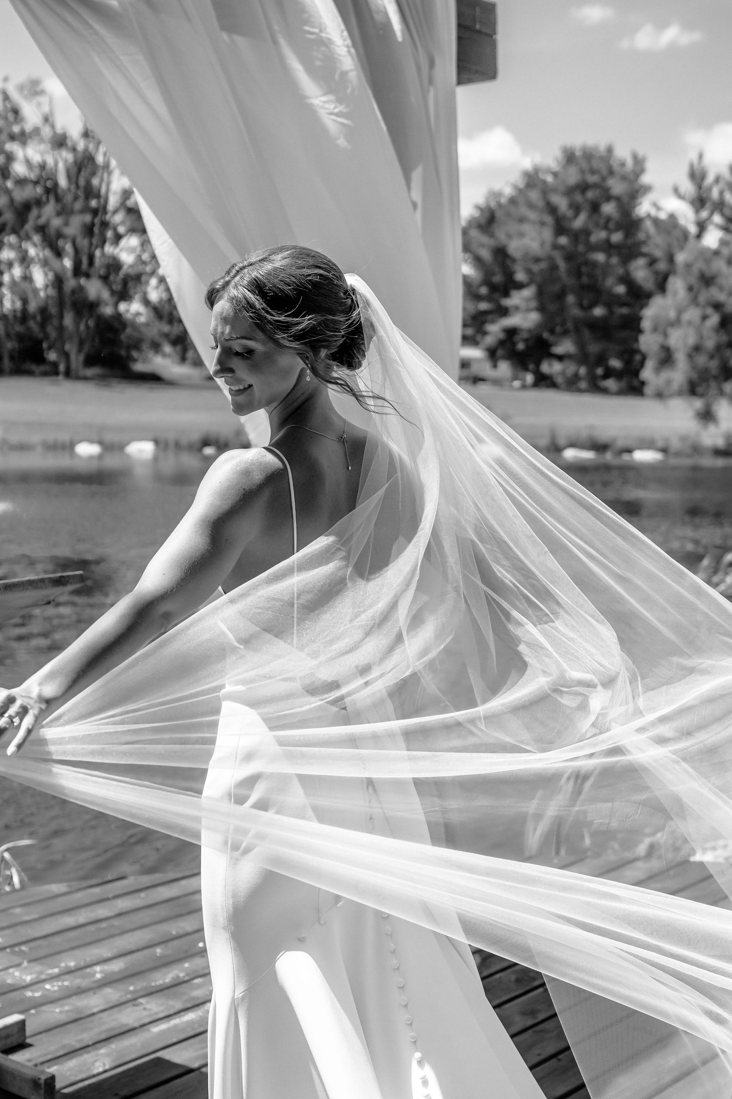 Black and white photo of a bride in a flowing gown with veil billowing in the wind near a lakeside dock.
