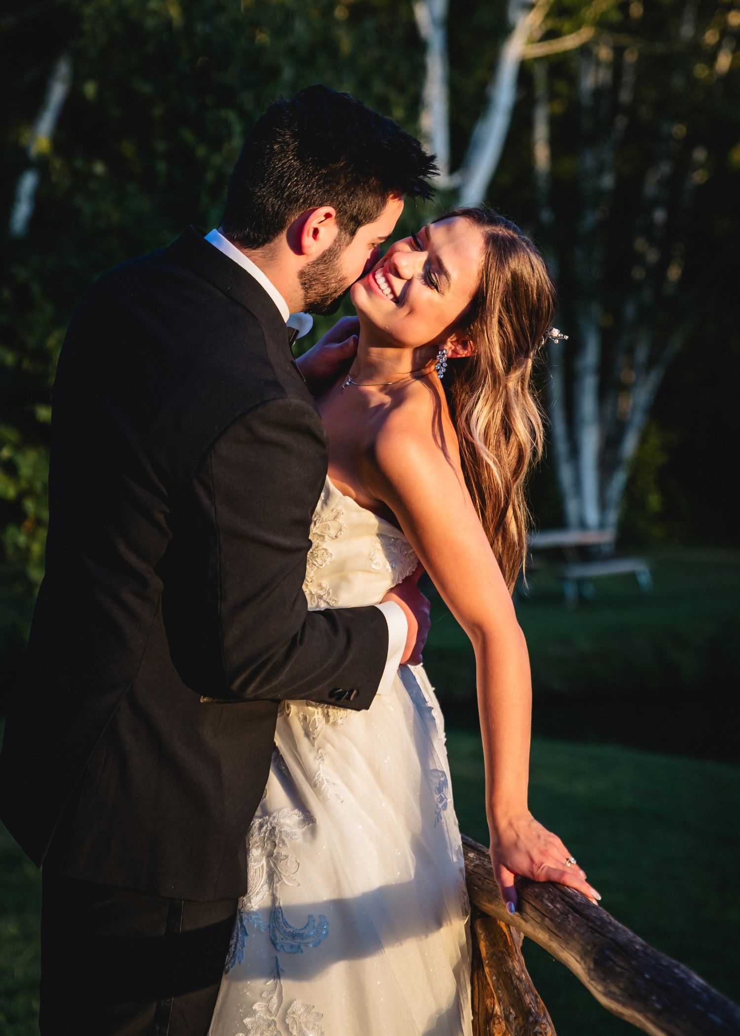 A smiling bride in a white lace gown embraces a groom in a black suit outdoors during golden hour.
