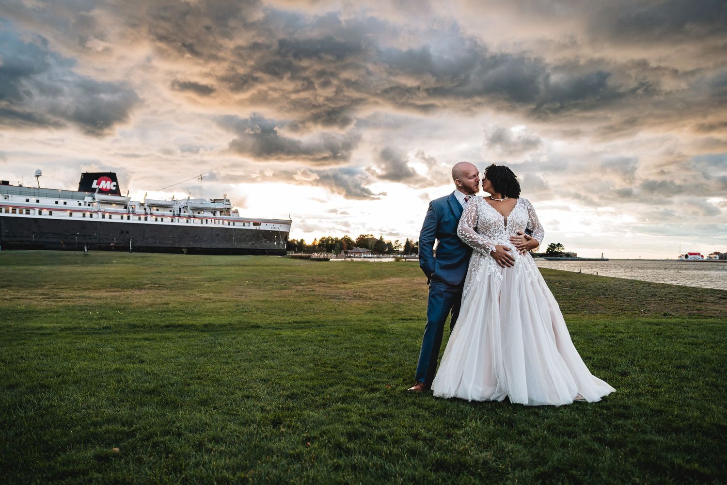 A newlywed couple shares a romantic moment in a grassy field under dramatic cloudy skies at sunset.