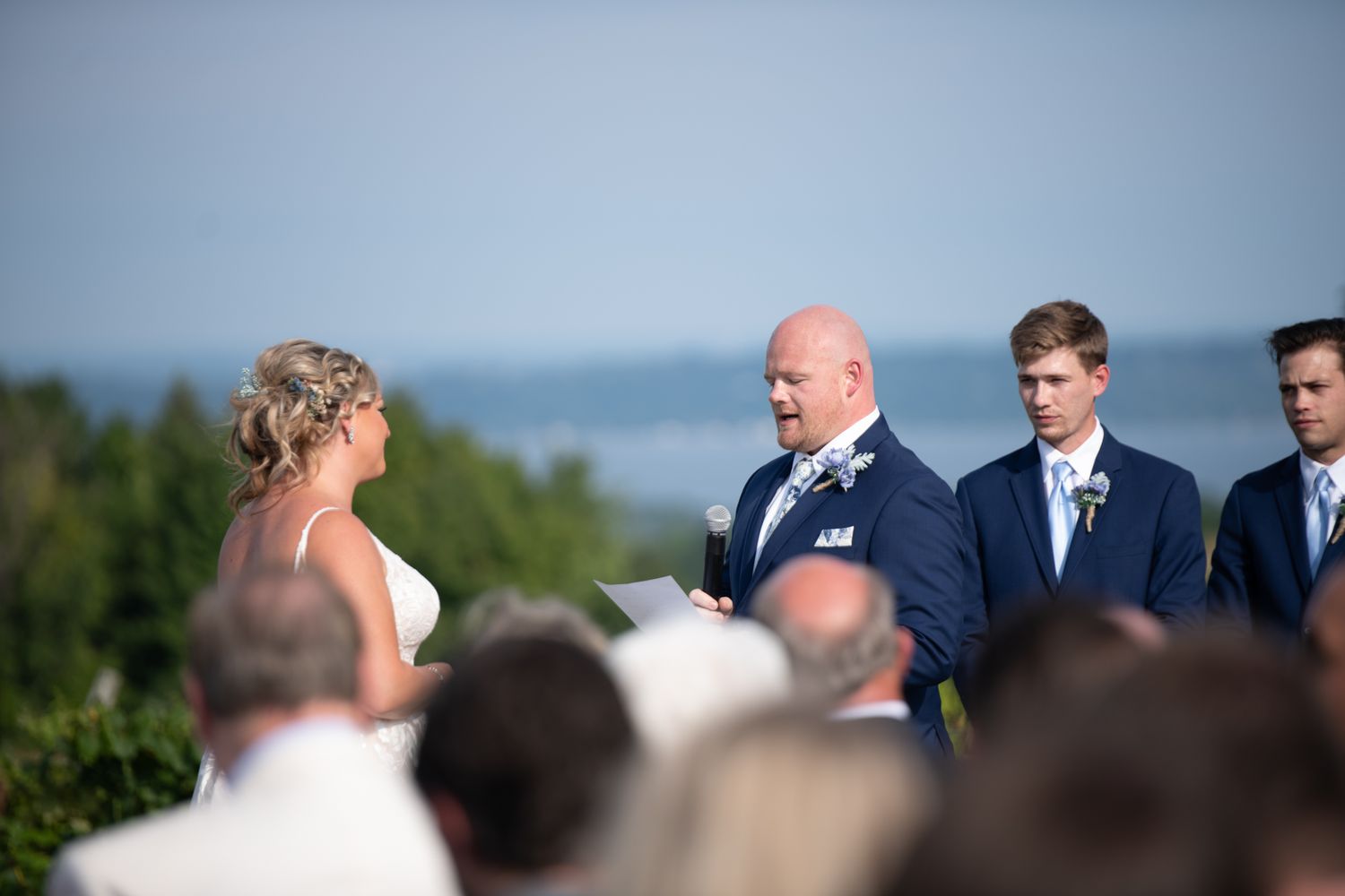 A group of people gather outdoors on a sunny day for a formal event with a scenic backdrop.