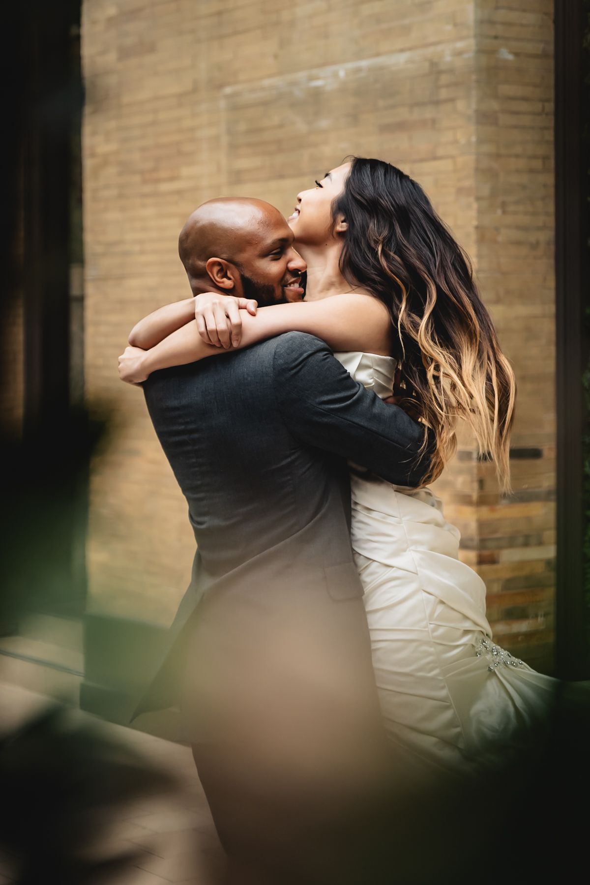 A couple embraces in an intimate dance pose against a brick wall backdrop with dramatic lighting.