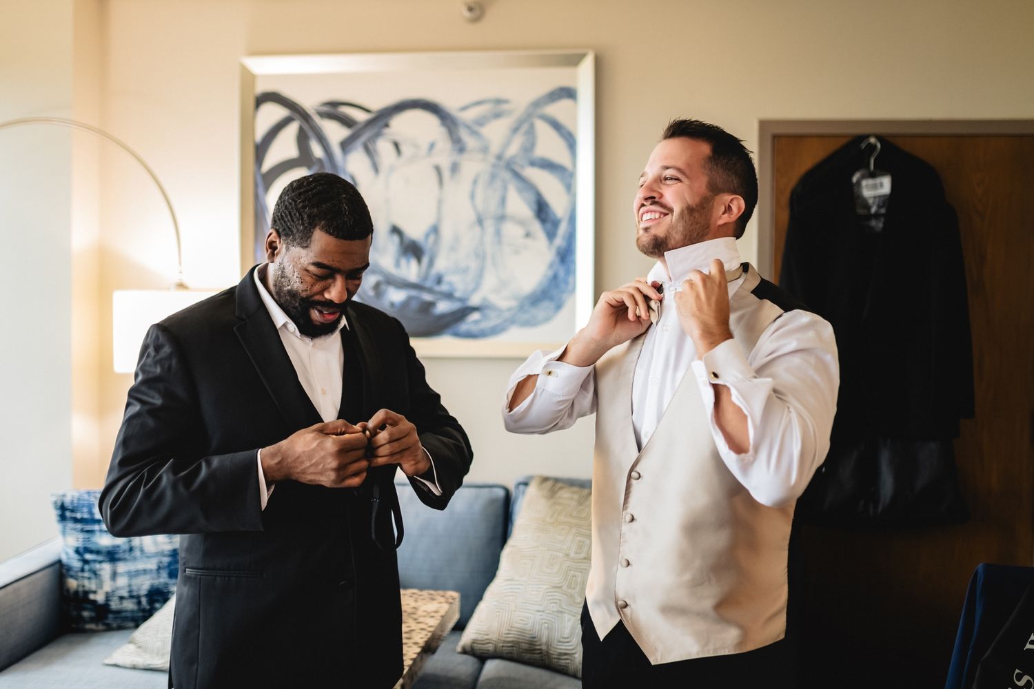 Two men in formal attire getting dressed in a room with abstract art on the wall and a garment hanging.