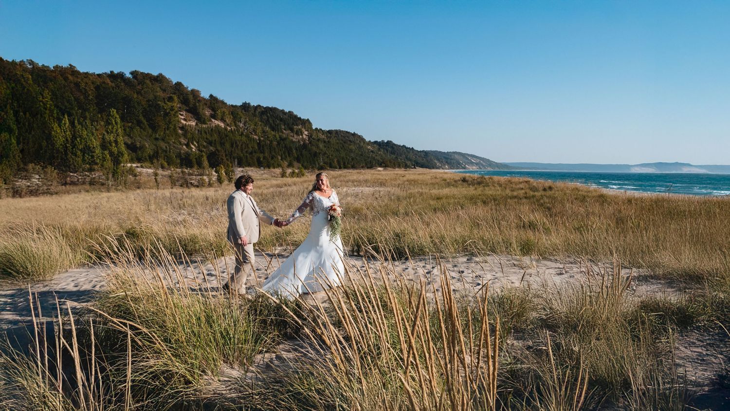 Bride and groom walking through beach dunes in Frankfort with Lake Michigan and forest backdrop on a wedding day at Lake Bluff Preserve