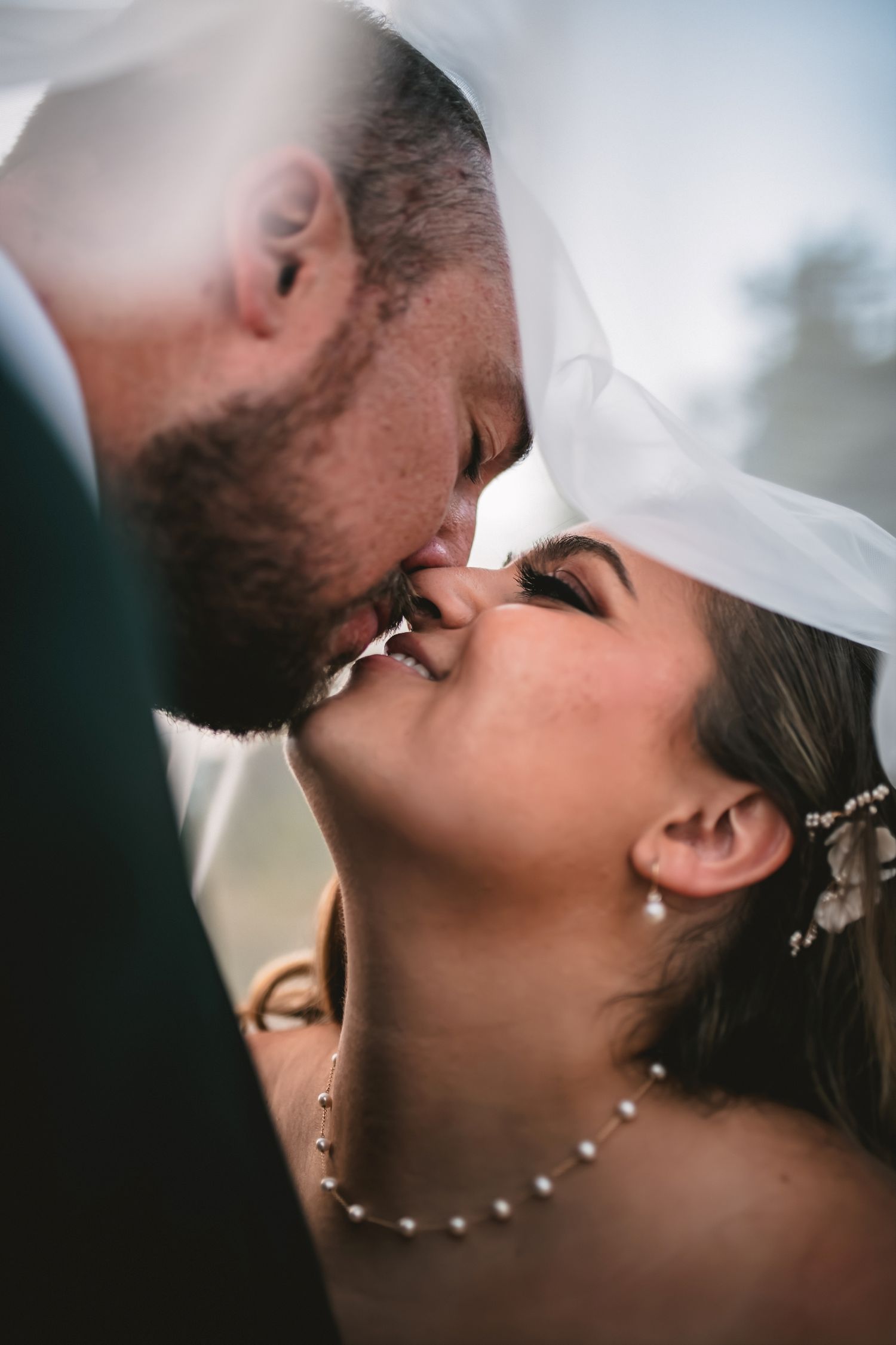 Romantic close-up wedding photo of newlyweds sharing an intimate kiss under a veil with delicate jewelry details.