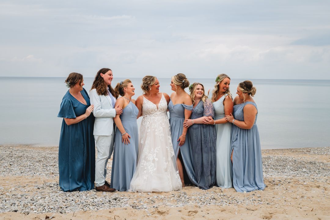 Bride in lace gown poses with bridesmaids in blue dresses on a rocky beach with ocean backdrop.