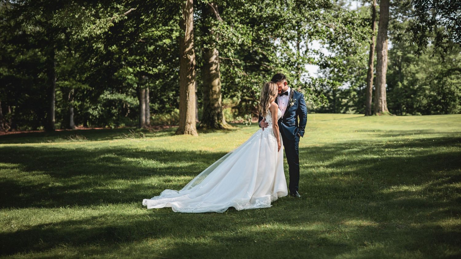A bride and groom share a romantic embrace on a lush green lawn surrounded by tall trees on their wedding day.