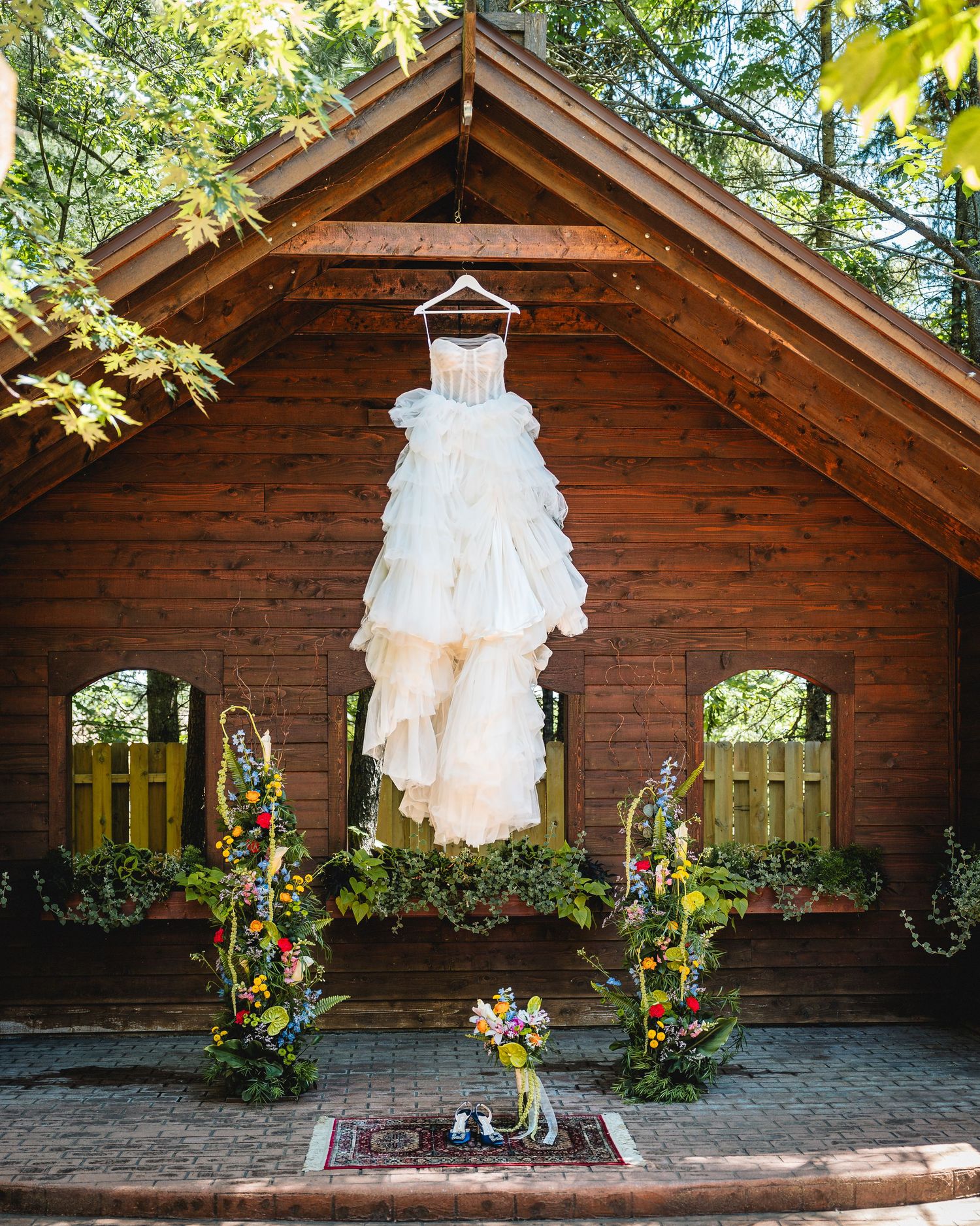 Elegant white wedding dress hanging on rustic wooden chapel exterior, surrounded by colorful floral arrangements and lush greenery.