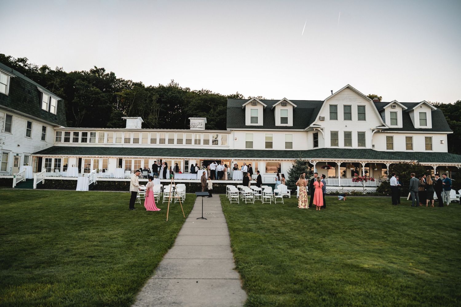 Guests gather on the lawn outside a large white colonial-style resort building with dormers during an outdoor event.