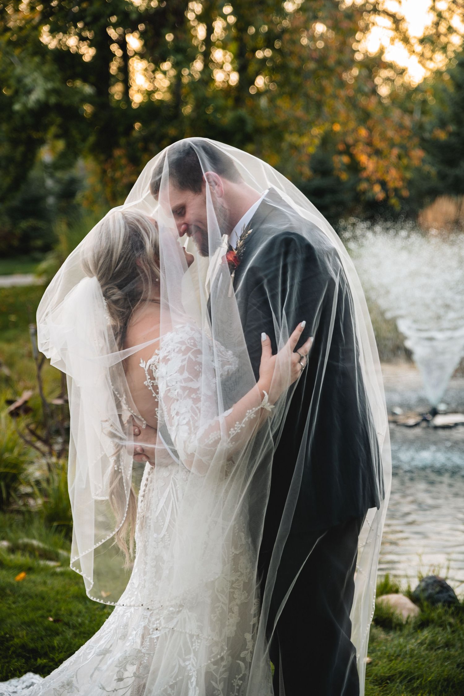 Bride and groom sharing a romantic kiss under a flowing wedding veil outdoors near a pond surrounded by autumn trees.