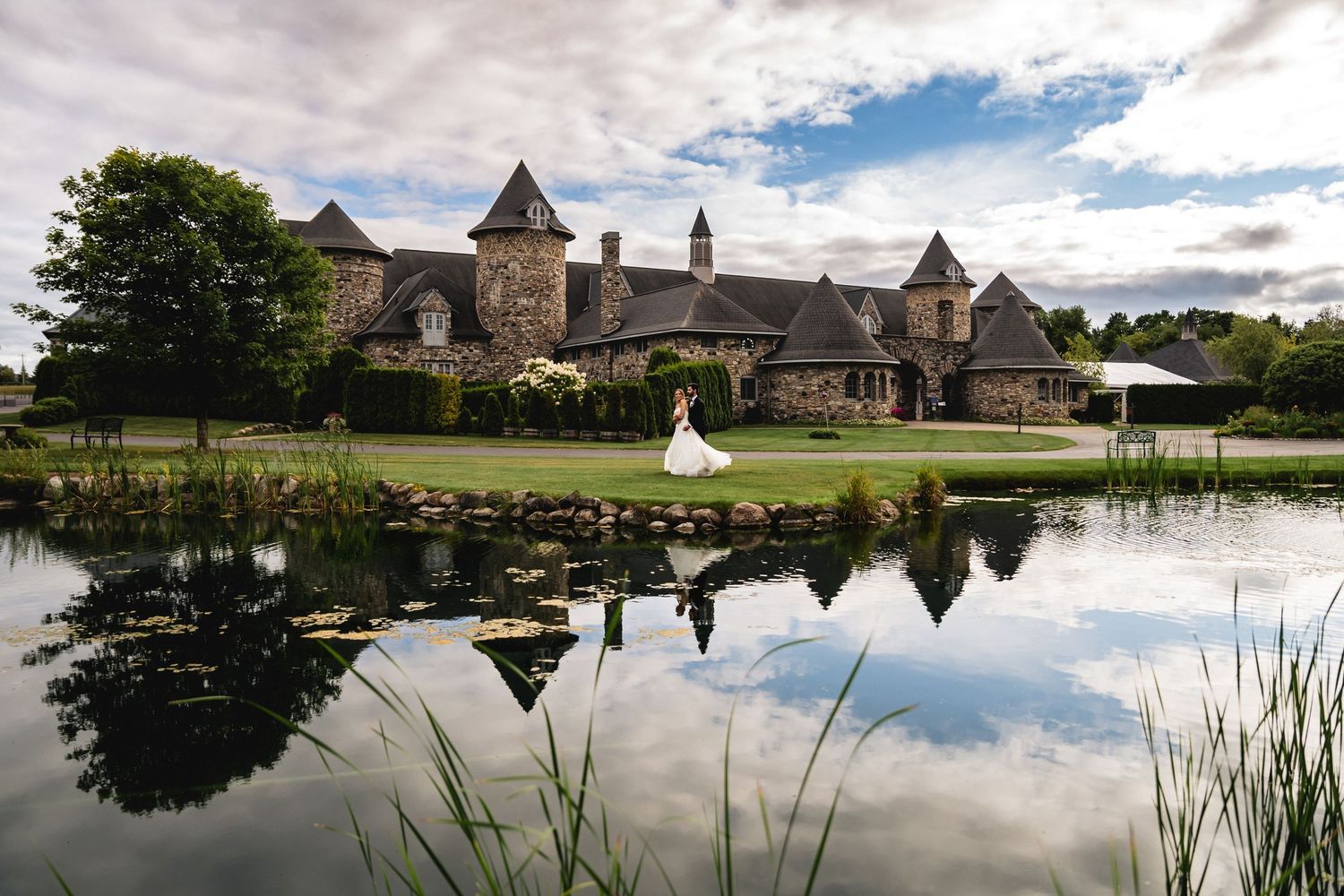 Scenic panoramic view of a rustic wedding venue with a lake, stone buildings, and lush green grounds under cloudy skies.