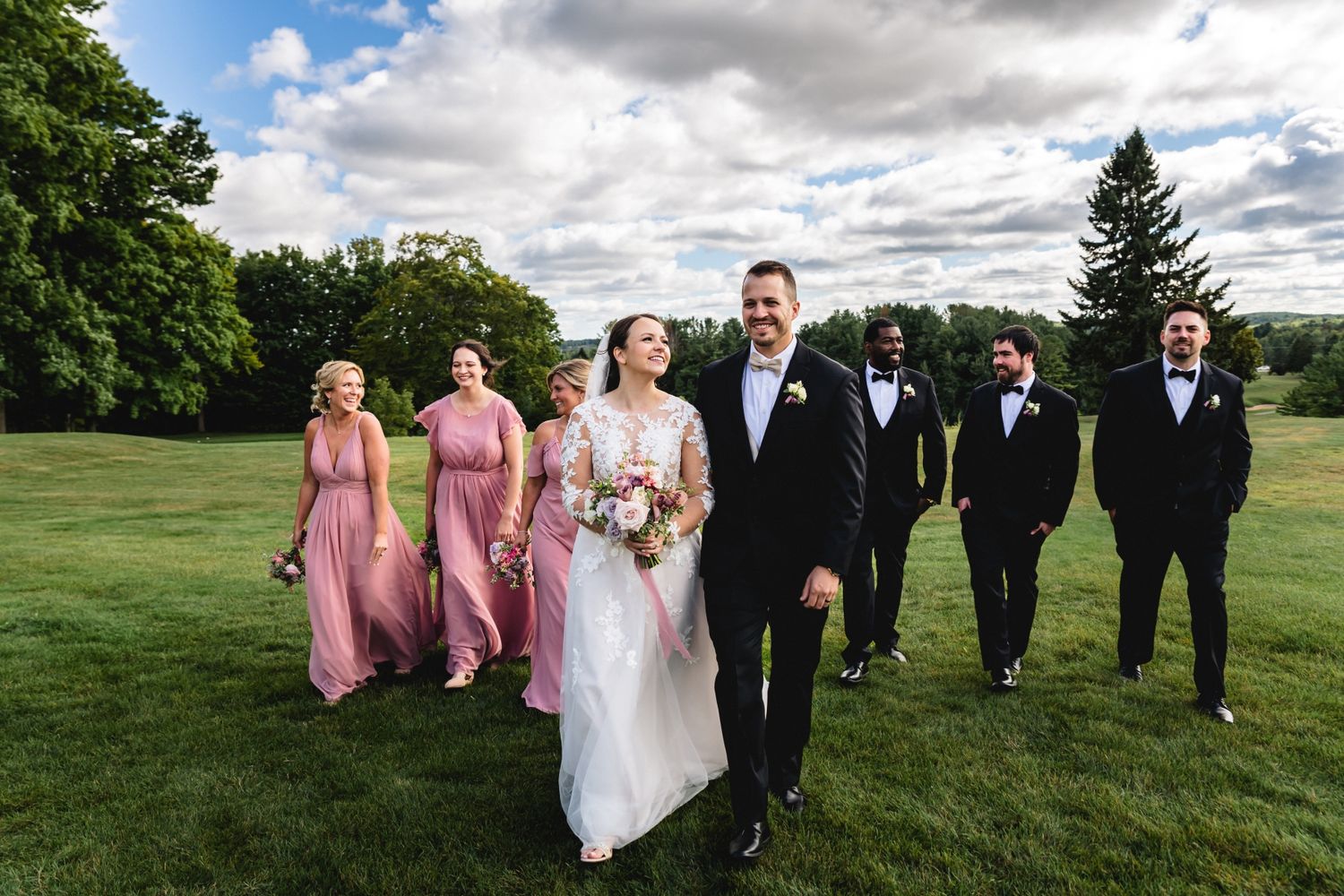 Wedding party members walk together across a lush green lawn under dramatic cloudy skies during an outdoor celebration.