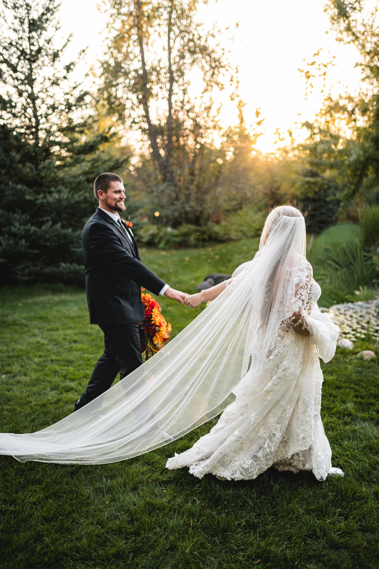 A couple in wedding attire twirls gracefully on a grassy lawn at sunset, with the bride's lace dress and veil flowing elegantly.