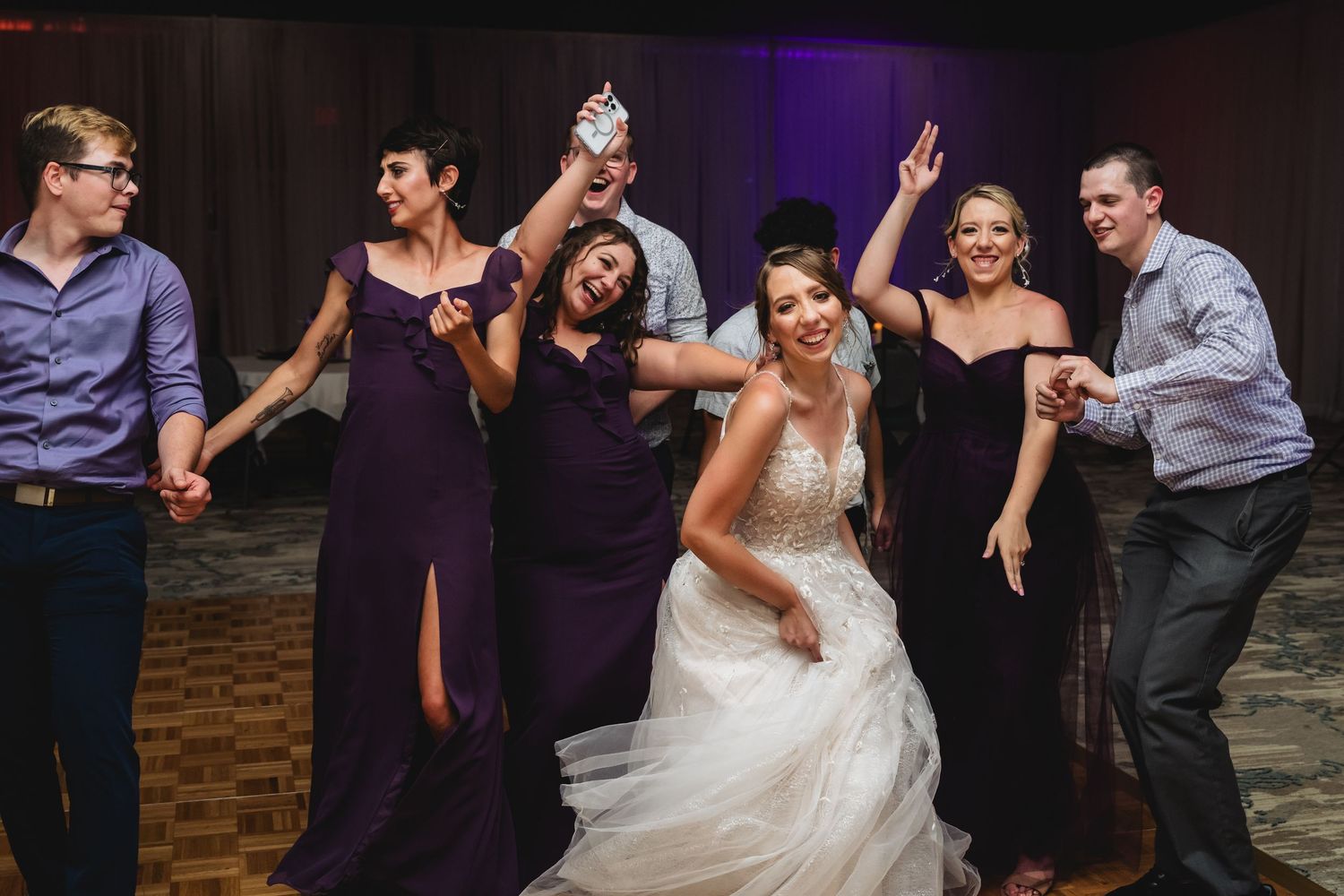 Bride in white gown dancing joyfully with bridesmaids in purple dresses at a lively wedding reception.