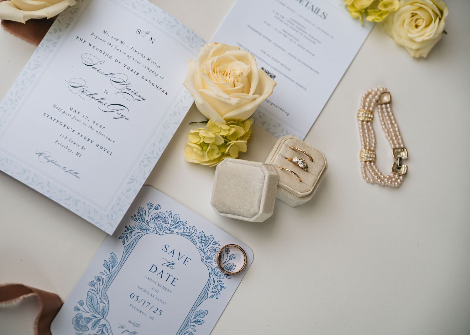 Elegant wedding flat lay with blue stationery, ring box, pearl bracelet, gold rings, and ivory roses on white background.