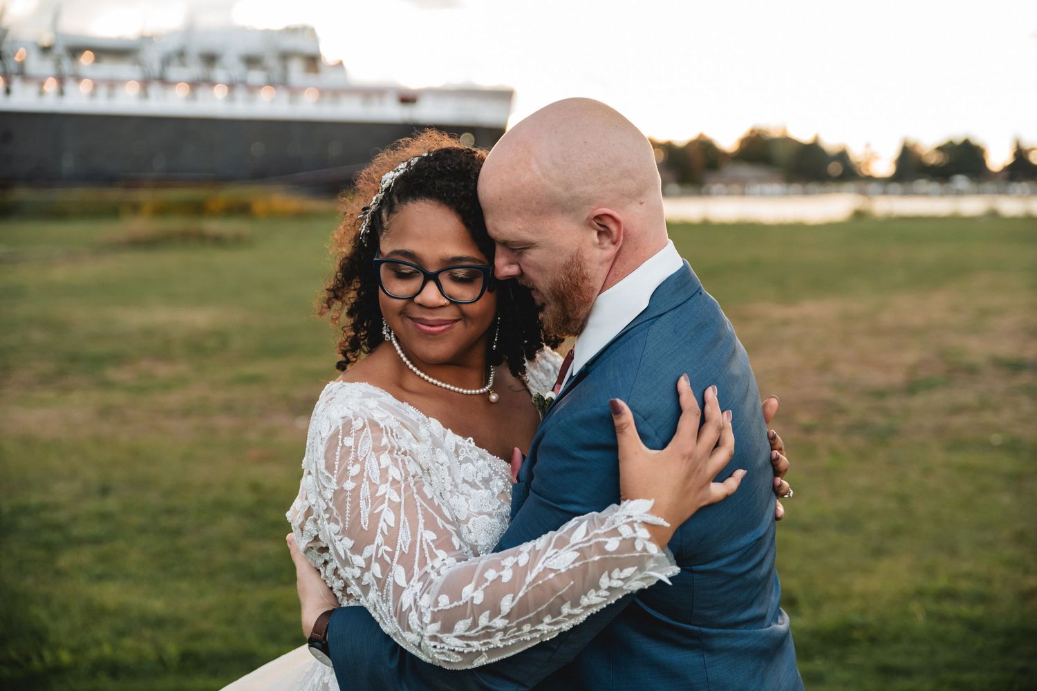 Interracial couple embracing outdoors in wedding attire, bride in lace gown with glasses, groom in blue suit.