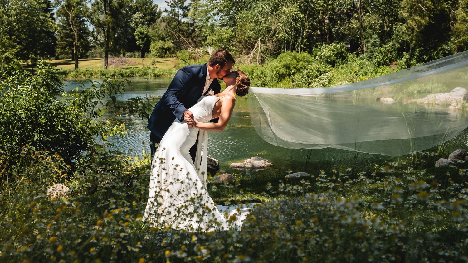 Bride and groom share a romantic kiss outdoors near a pond, with her floral gown and flowing veil billowing in the breeze.