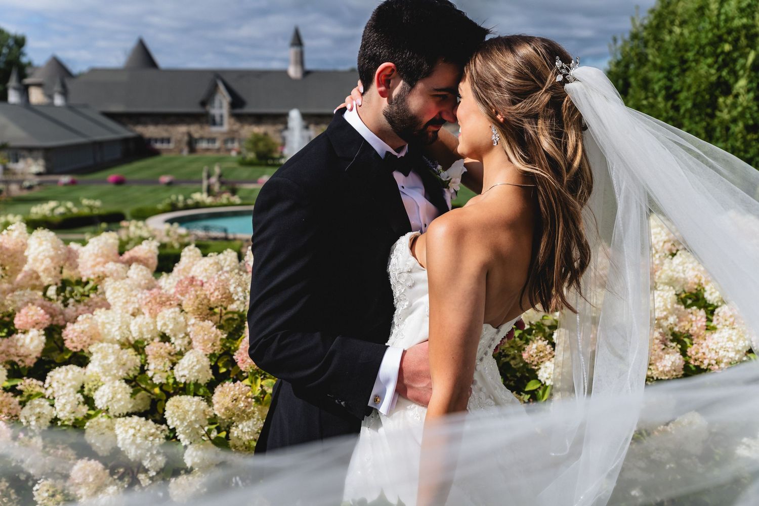 Bride and groom share romantic moment surrounded by white hydrangeas, veil flowing, with elegant estate gardens in background.