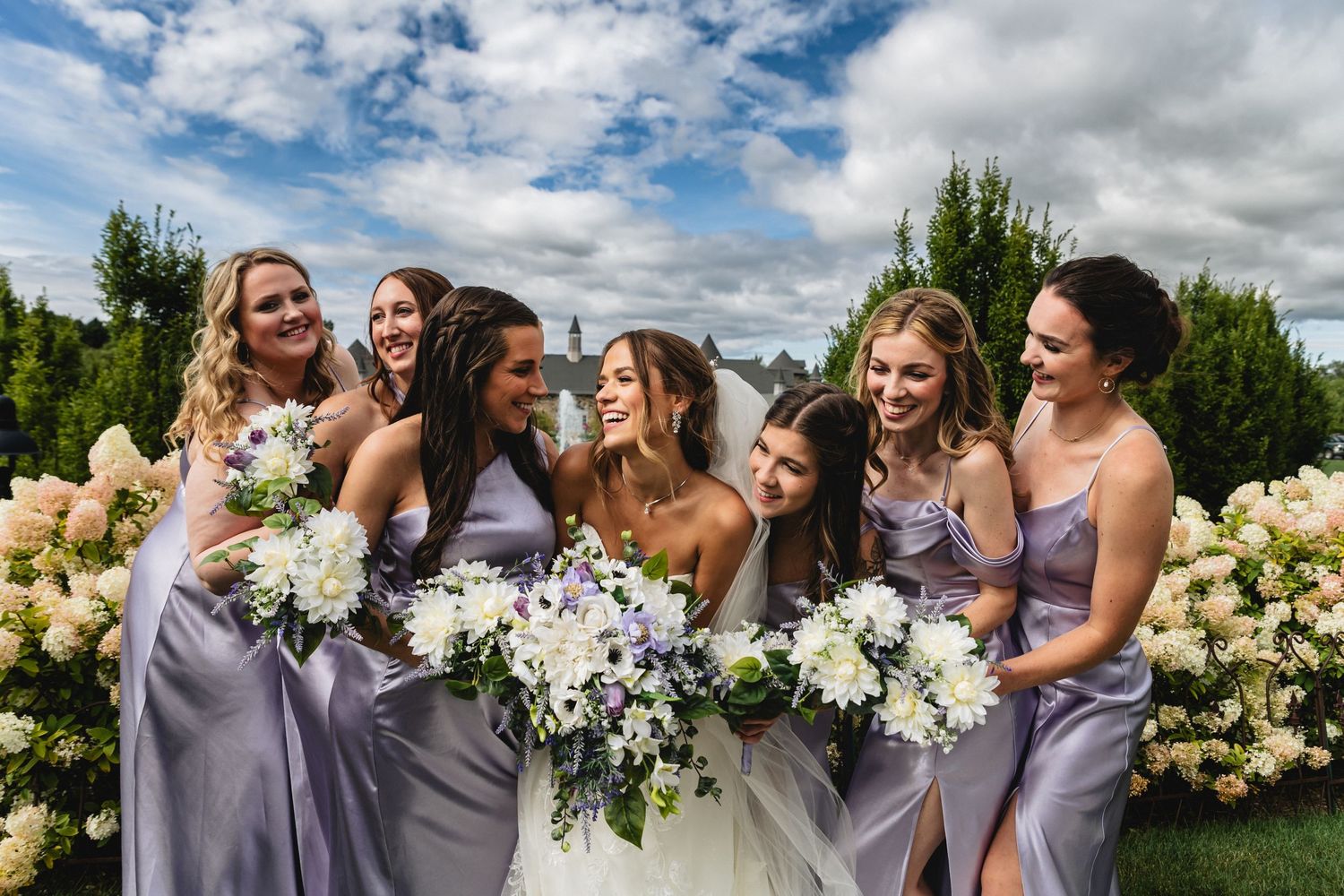 Wedding party wearing lavender bridesmaid dresses poses together outdoors under blue sky with white clouds.