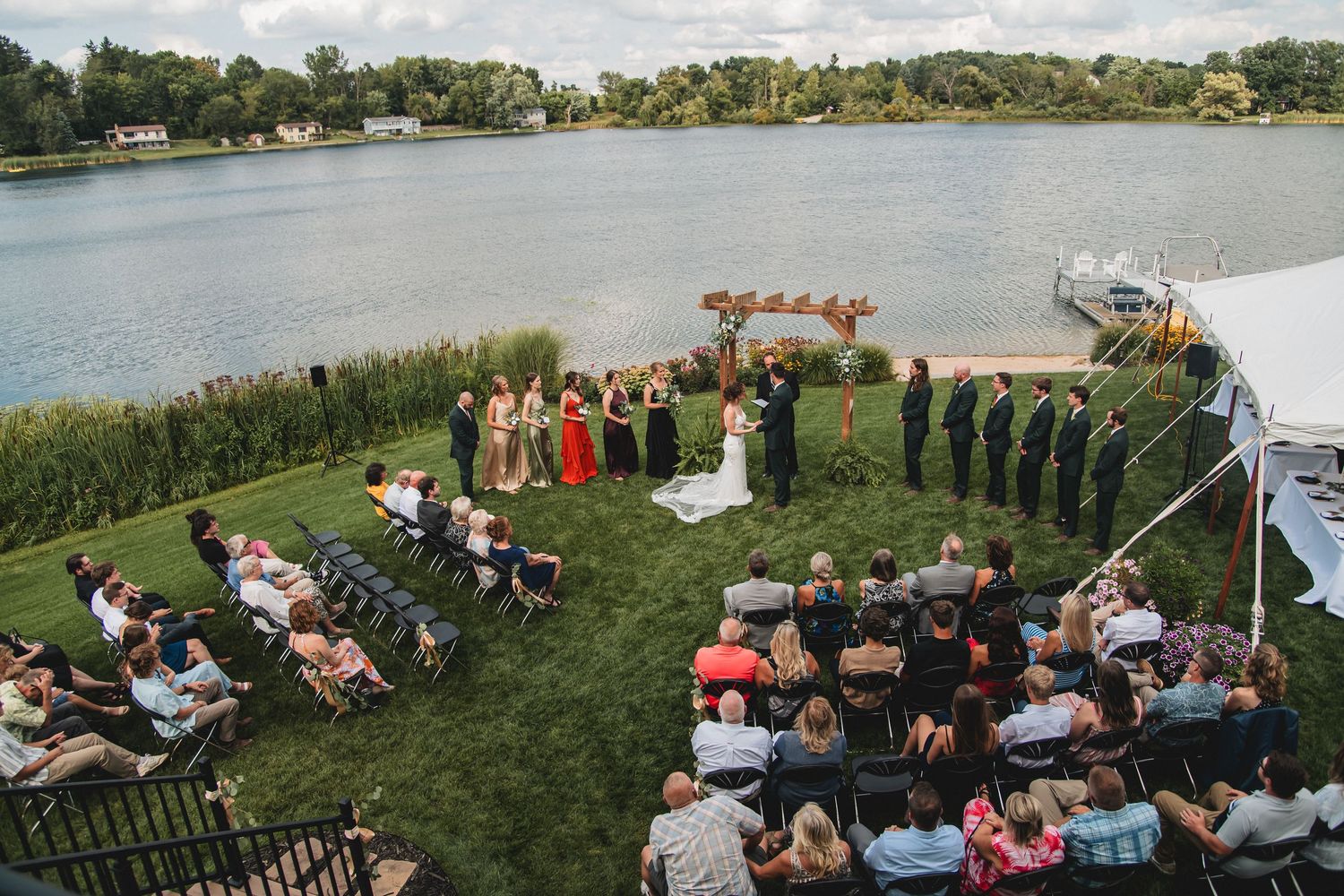 Outdoor lakeside wedding ceremony with wedding party and guests seated on green lawn with water and trees in background.